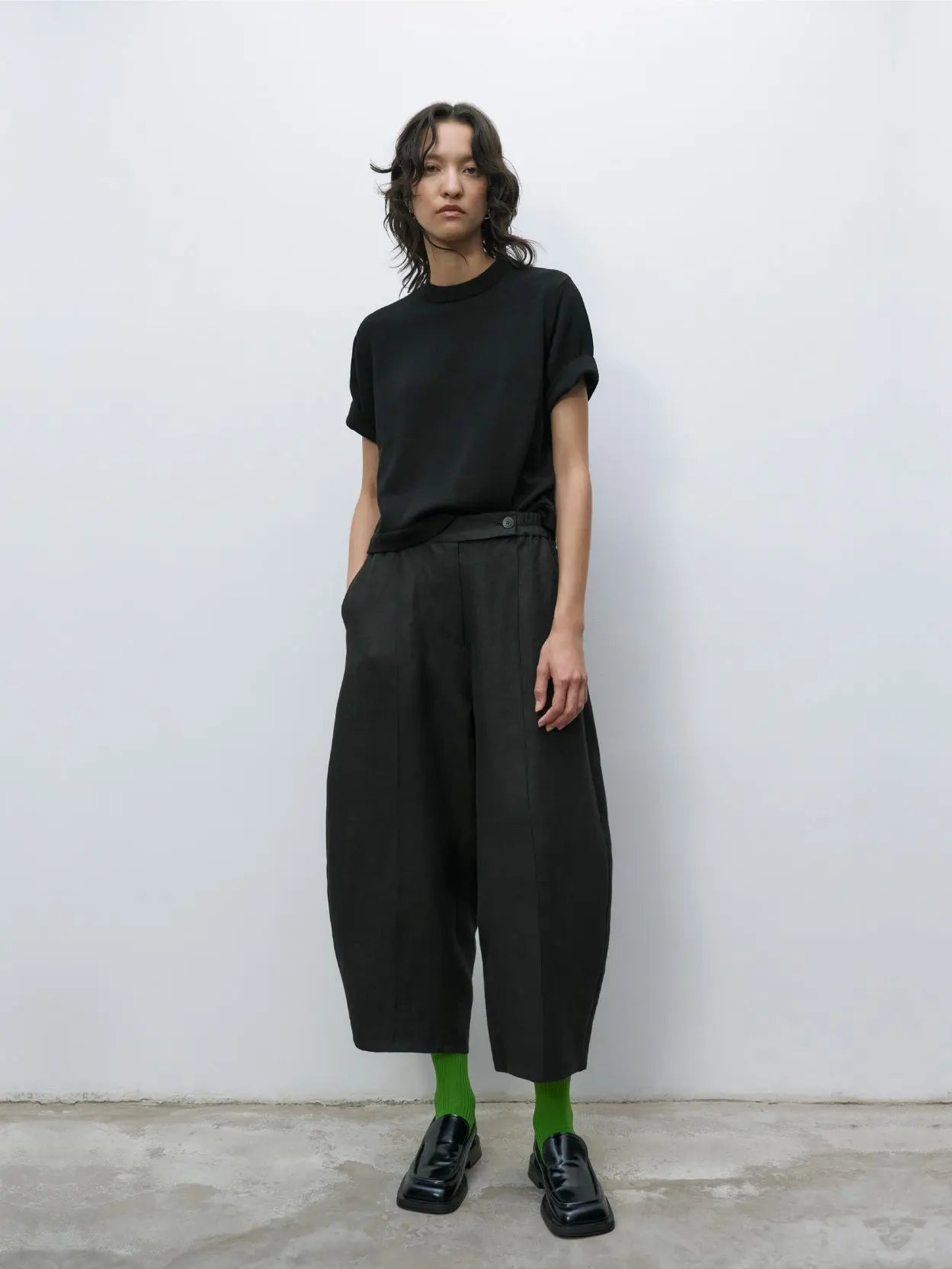 A pair of black, wide-leg trousers with an elastic waistband and a single button closure. The trousers have a clean, minimalist design with visible front pleats running from the waistband to the hem. The fabric appears to be smooth and slightly structured, available exclusively at our Barcelona store. These are the Linen Curved Pants Black by Cordera.