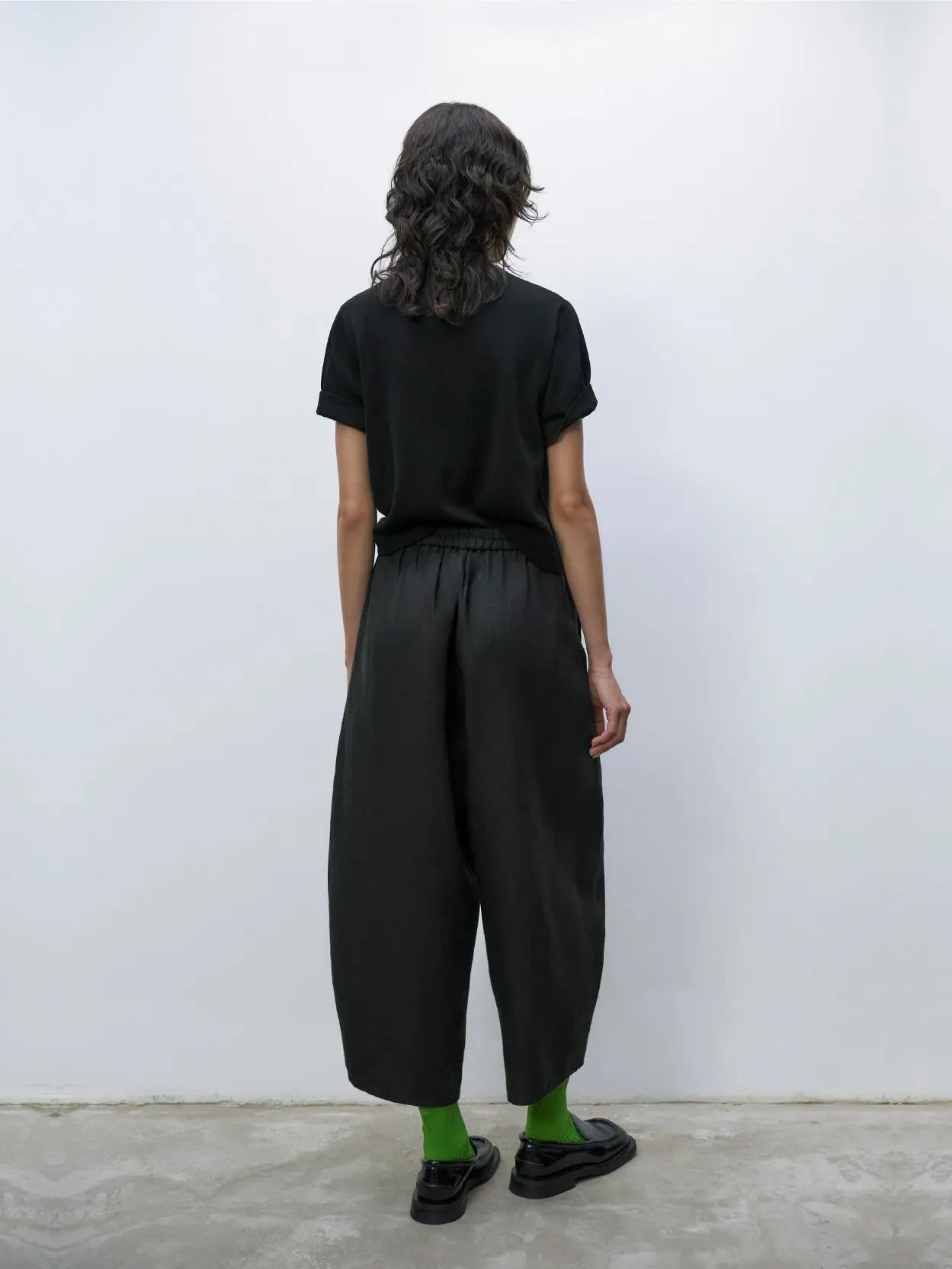 A pair of black, wide-leg trousers with an elastic waistband and a single button closure. The trousers have a clean, minimalist design with visible front pleats running from the waistband to the hem. The fabric appears to be smooth and slightly structured, available exclusively at our Barcelona store. These are the Linen Curved Pants Black by Cordera.