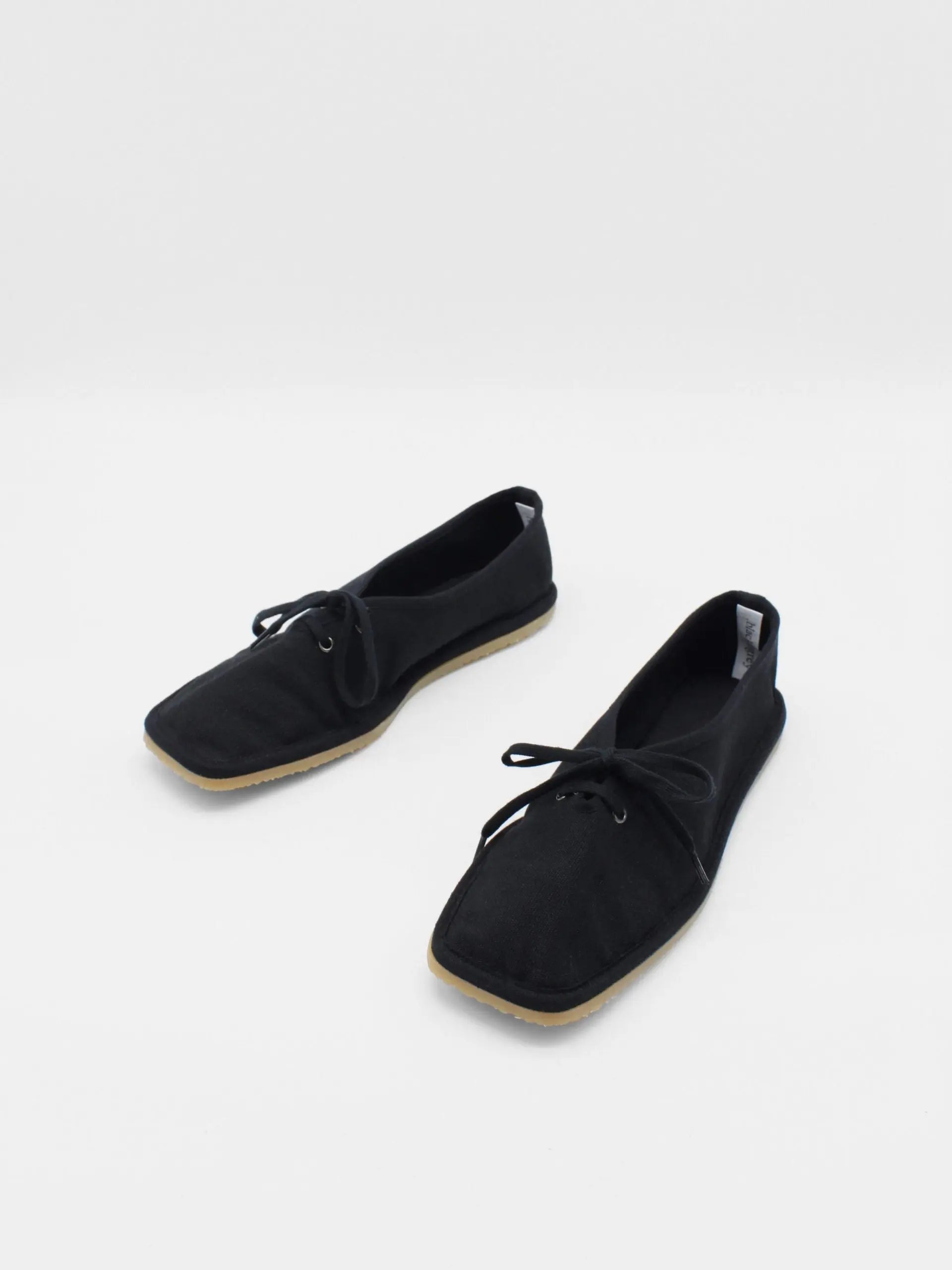 A pair of Lona Black shoes by About Arianne with a minimalist, square-toe design, positioned side by side on a white background. The shoes have black laces and appear to be made of a fabric material, reflecting the chic style found in Barcelona's finest stores.