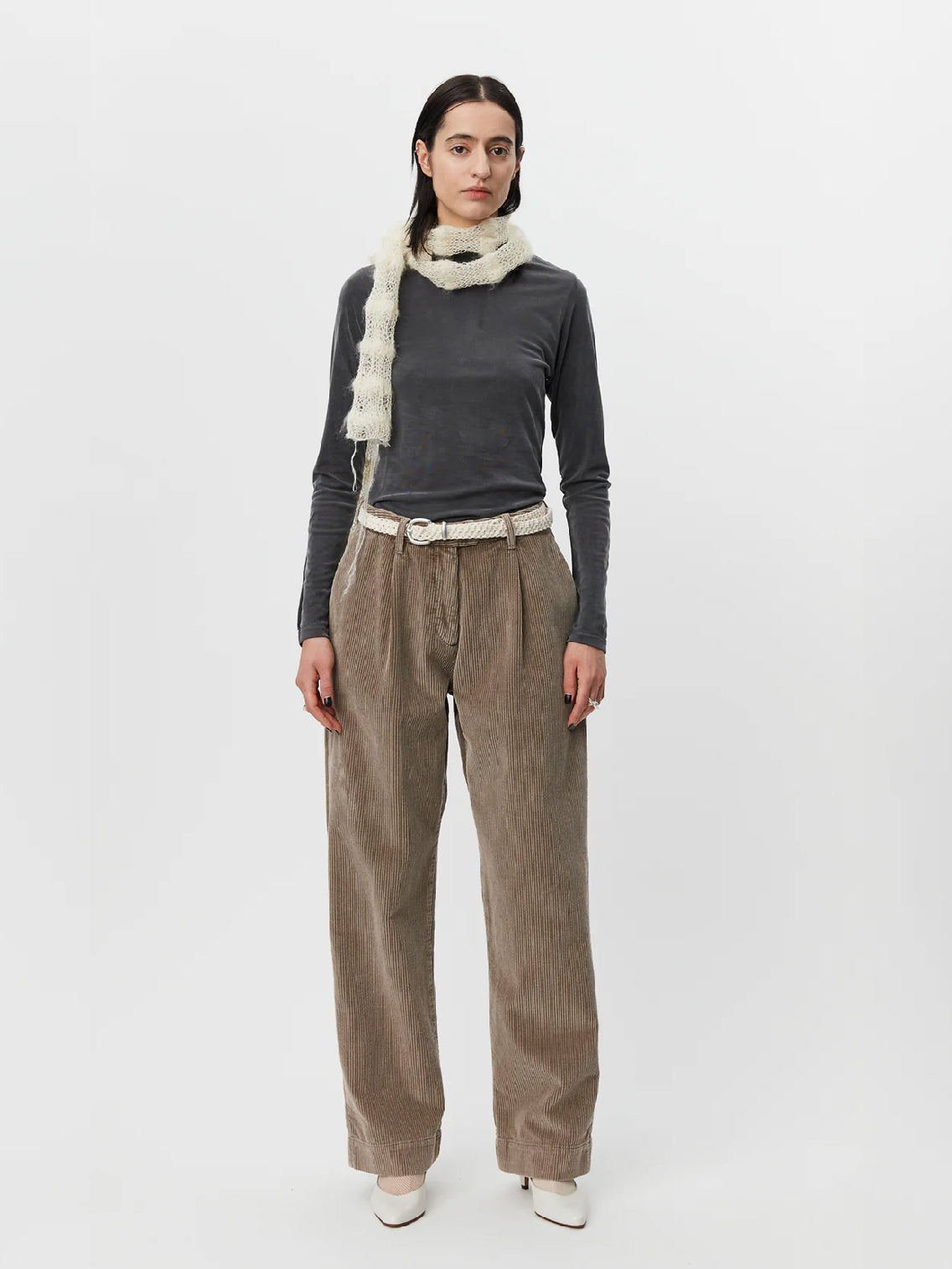 A person with long dark hair wears a dark gray long-sleeve top paired with beige high-waisted Mfpen Corduroy Trousers in Dusty Brown. They have a white knit scarf loosely draped around their neck and a white belt. The outfit is completed with white heeled shoes, all set against a plain white background.