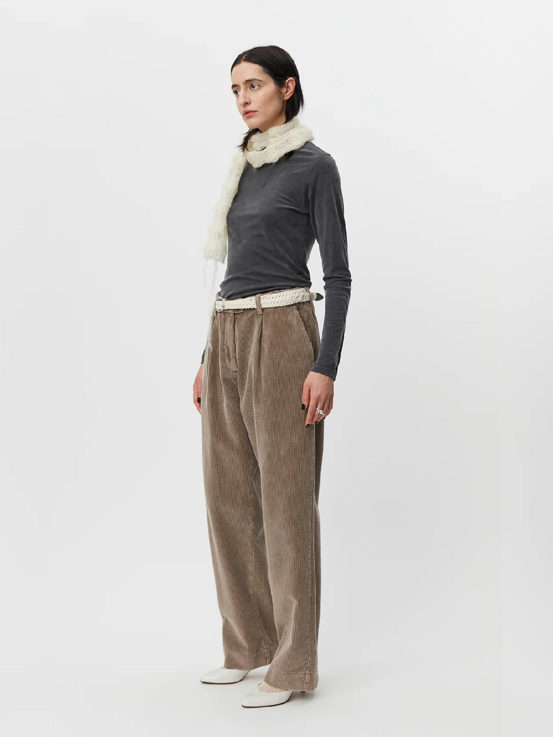 A person with long dark hair wears a dark gray long-sleeve top paired with beige high-waisted Mfpen Corduroy Trousers in Dusty Brown. They have a white knit scarf loosely draped around their neck and a white belt. The outfit is completed with white heeled shoes, all set against a plain white background.