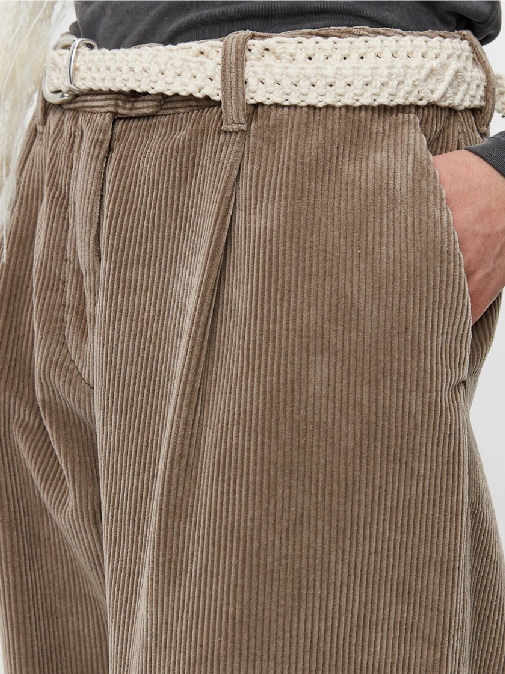 A person with long dark hair wears a dark gray long-sleeve top paired with beige high-waisted Mfpen Corduroy Trousers in Dusty Brown. They have a white knit scarf loosely draped around their neck and a white belt. The outfit is completed with white heeled shoes, all set against a plain white background.