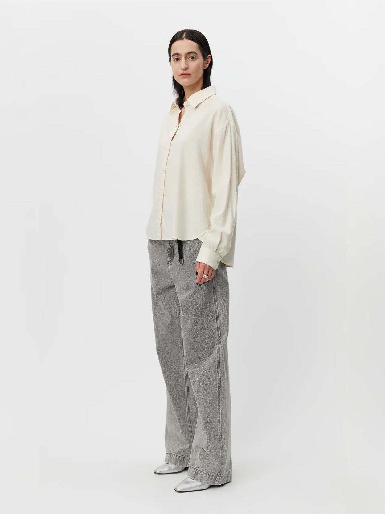 A person with long dark hair stands against a plain white background. They are wearing a Peeled Ivory Exec Shirt by Mfpen and gray wide-leg jeans, with hands relaxed by their sides. The jeans have a loose, baggy fit, and the person is wearing silver shoes.