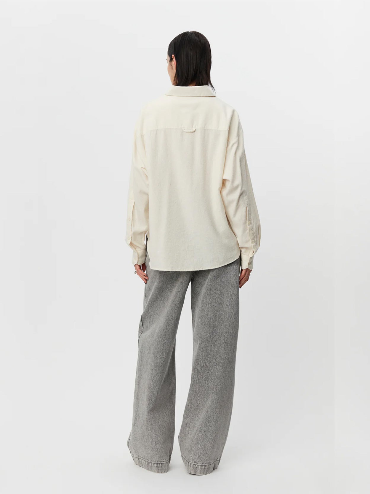 A person with long dark hair stands against a plain white background. They are wearing a Peeled Ivory Exec Shirt by Mfpen and gray wide-leg jeans, with hands relaxed by their sides. The jeans have a loose, baggy fit, and the person is wearing silver shoes.