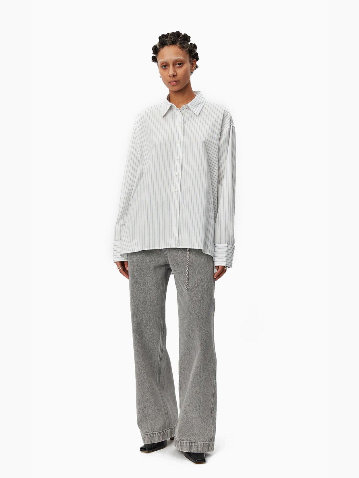 A person with short, braided hair wears an oversized-fit Mfpen Irregular Shirt Blue Stripe, made of deadstock cotton, and wide-legged gray trousers. The shirt features vertical stripes and an irregular hem. They stand against a plain white background, looking forward with a neutral expression.