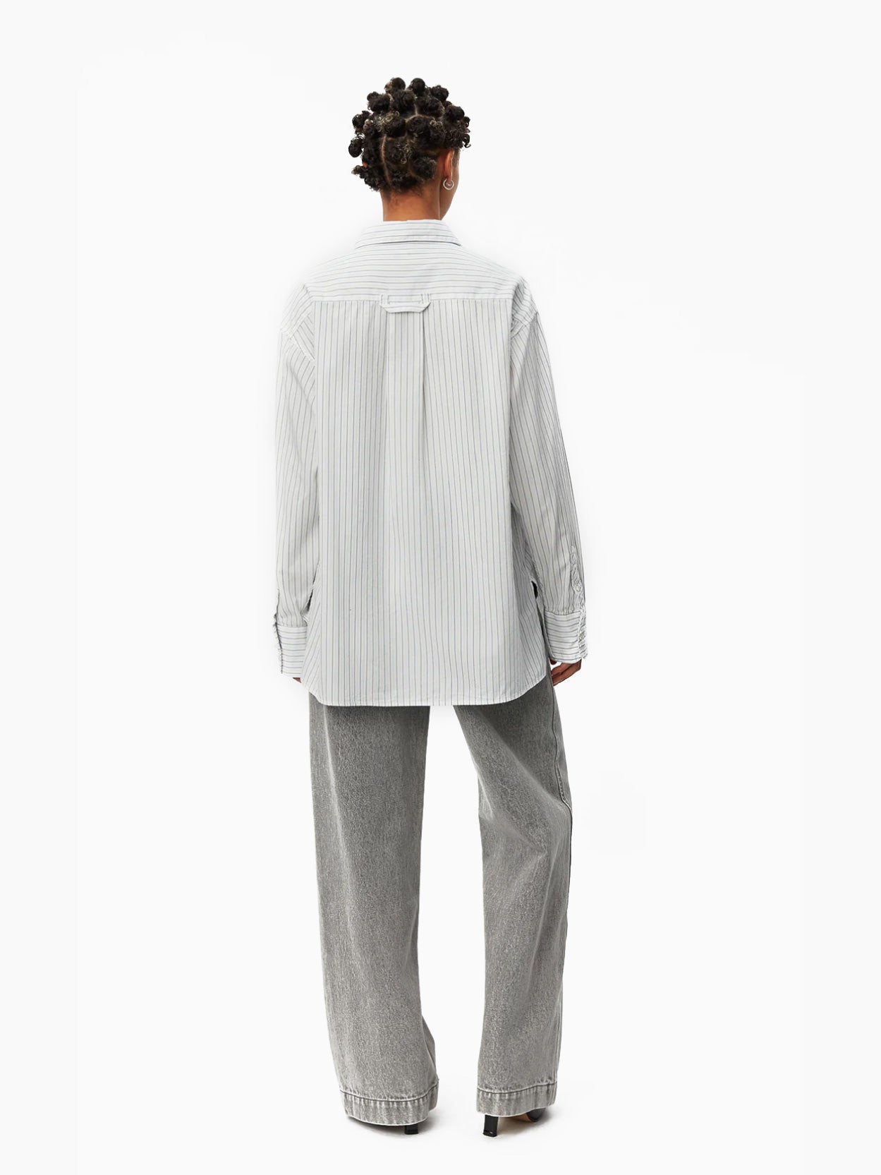 A person with short, braided hair wears an oversized-fit Mfpen Irregular Shirt Blue Stripe, made of deadstock cotton, and wide-legged gray trousers. The shirt features vertical stripes and an irregular hem. They stand against a plain white background, looking forward with a neutral expression.