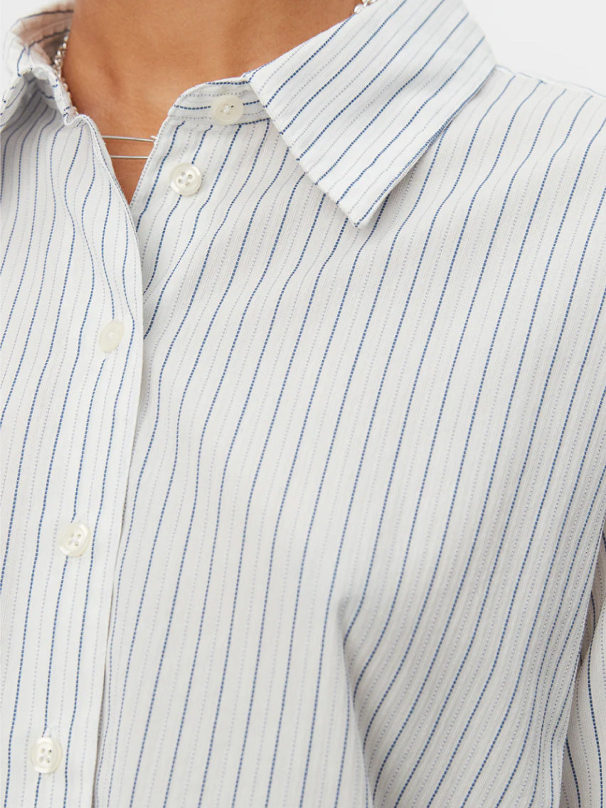 A person with short, braided hair wears an oversized-fit Mfpen Irregular Shirt Blue Stripe, made of deadstock cotton, and wide-legged gray trousers. The shirt features vertical stripes and an irregular hem. They stand against a plain white background, looking forward with a neutral expression.