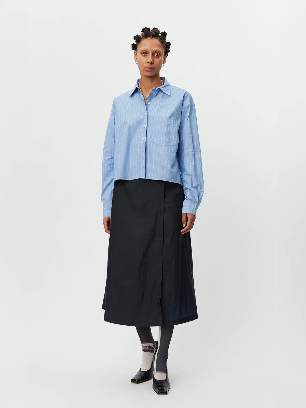 A person standing against a white background is wearing the New Format Shirt Light Blue by Mfpen, featuring long sleeves, a button-up design with a collar and button placket. They are paired with a black midi skirt, dark gray tights, and black shoes. The cotton poplin shirt complements their short, curly hair styled in twists.