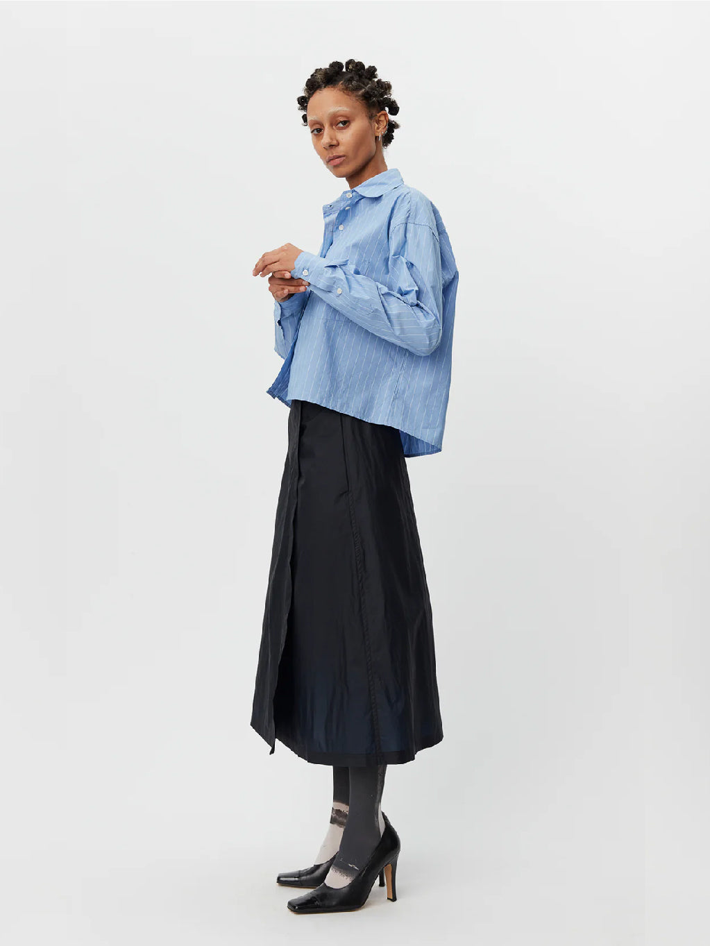 A person standing against a white background is wearing the New Format Shirt Light Blue by Mfpen, featuring long sleeves, a button-up design with a collar and button placket. They are paired with a black midi skirt, dark gray tights, and black shoes. The cotton poplin shirt complements their short, curly hair styled in twists.