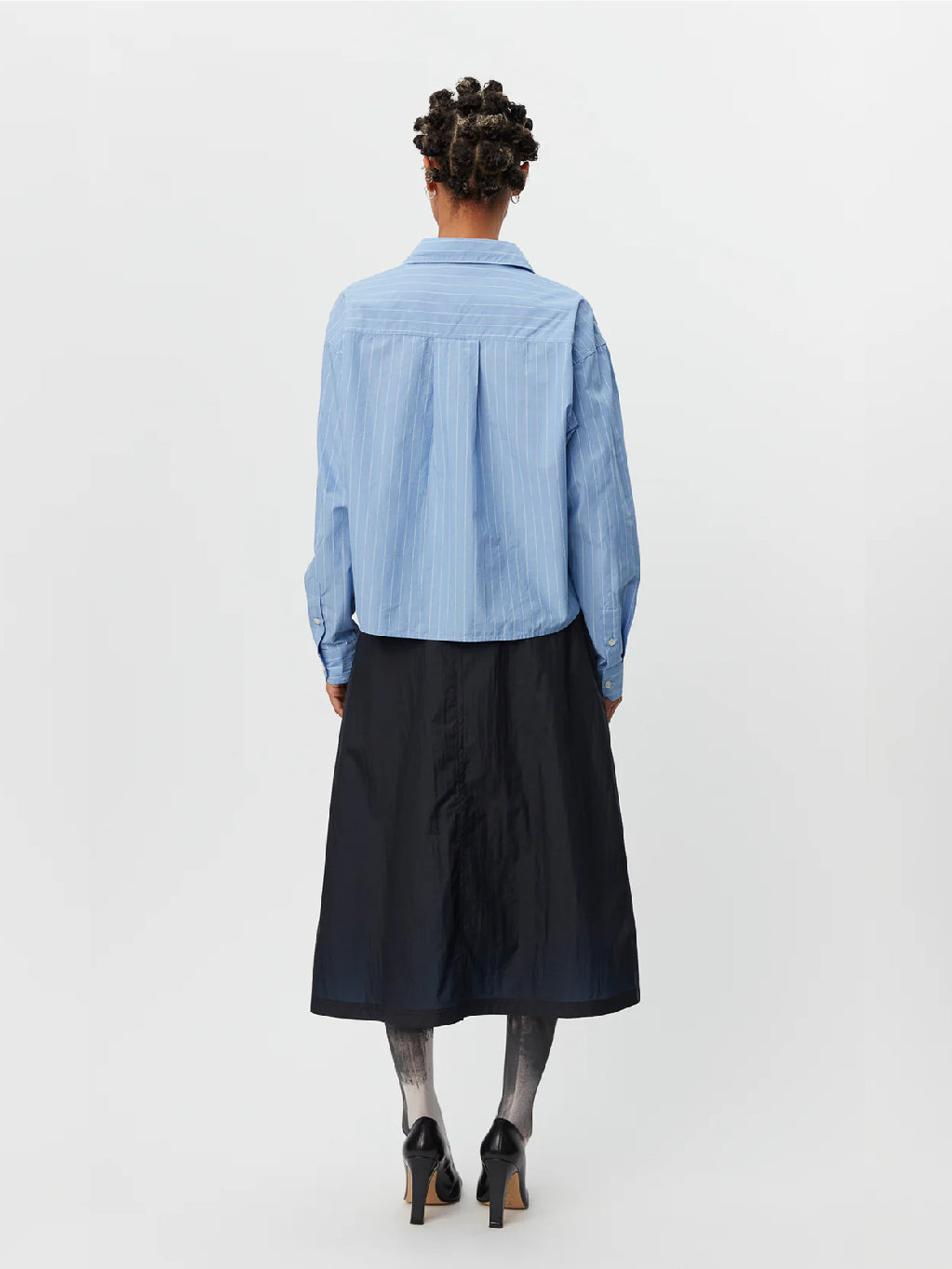 A person standing against a white background is wearing the New Format Shirt Light Blue by Mfpen, featuring long sleeves, a button-up design with a collar and button placket. They are paired with a black midi skirt, dark gray tights, and black shoes. The cotton poplin shirt complements their short, curly hair styled in twists.