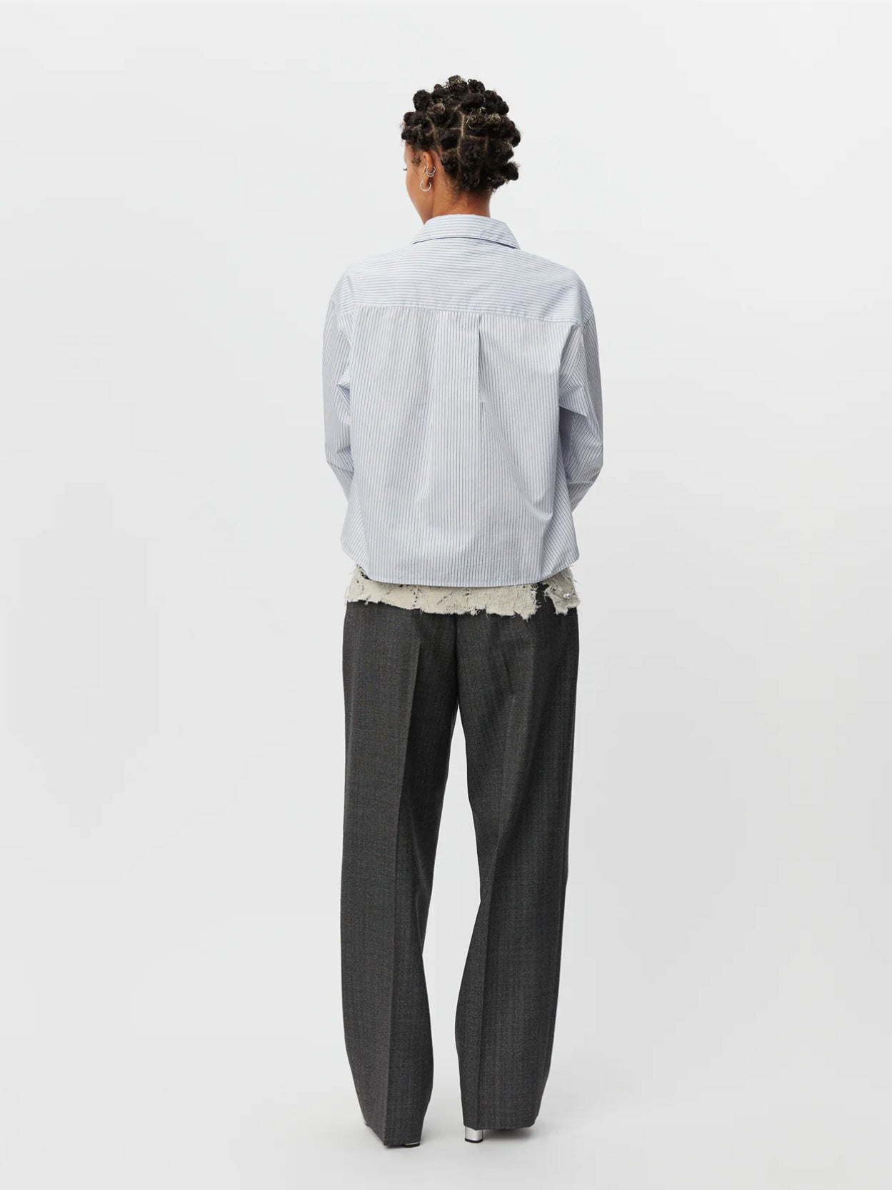 A person with short, styled hair stands against a plain white background. They are wearing the Mfpen New Format Shirt in Blue Stripe, an off-white lace top underneath, and loose-fitting gray trousers in a relaxed boxy fit, paired with silver pointed shoes. They have a neutral expression.