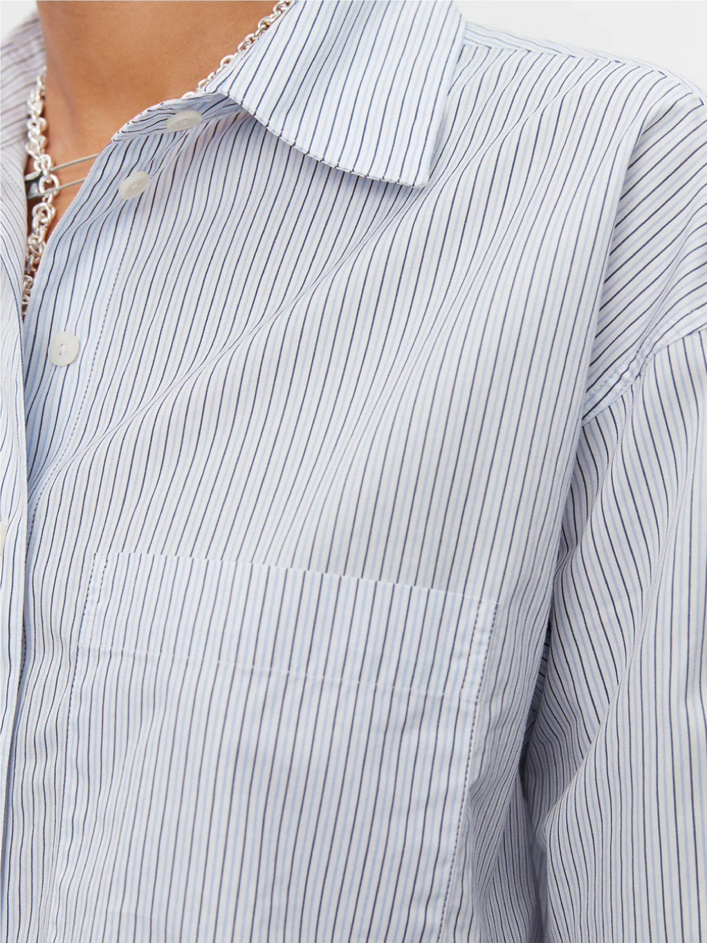 A person with short, styled hair stands against a plain white background. They are wearing the Mfpen New Format Shirt in Blue Stripe, an off-white lace top underneath, and loose-fitting gray trousers in a relaxed boxy fit, paired with silver pointed shoes. They have a neutral expression.