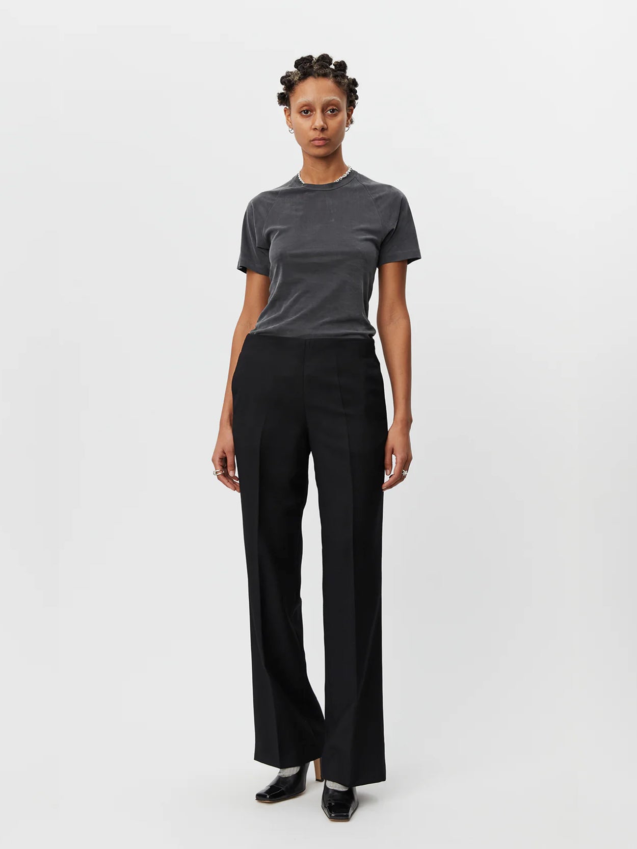 A person stands against a plain white background, wearing a fitted gray short-sleeve T-shirt and black Taper Trousers Black by Mfpen made from virgin wool. They have short hair styled in twists and are wearing black shoes with a small heel.