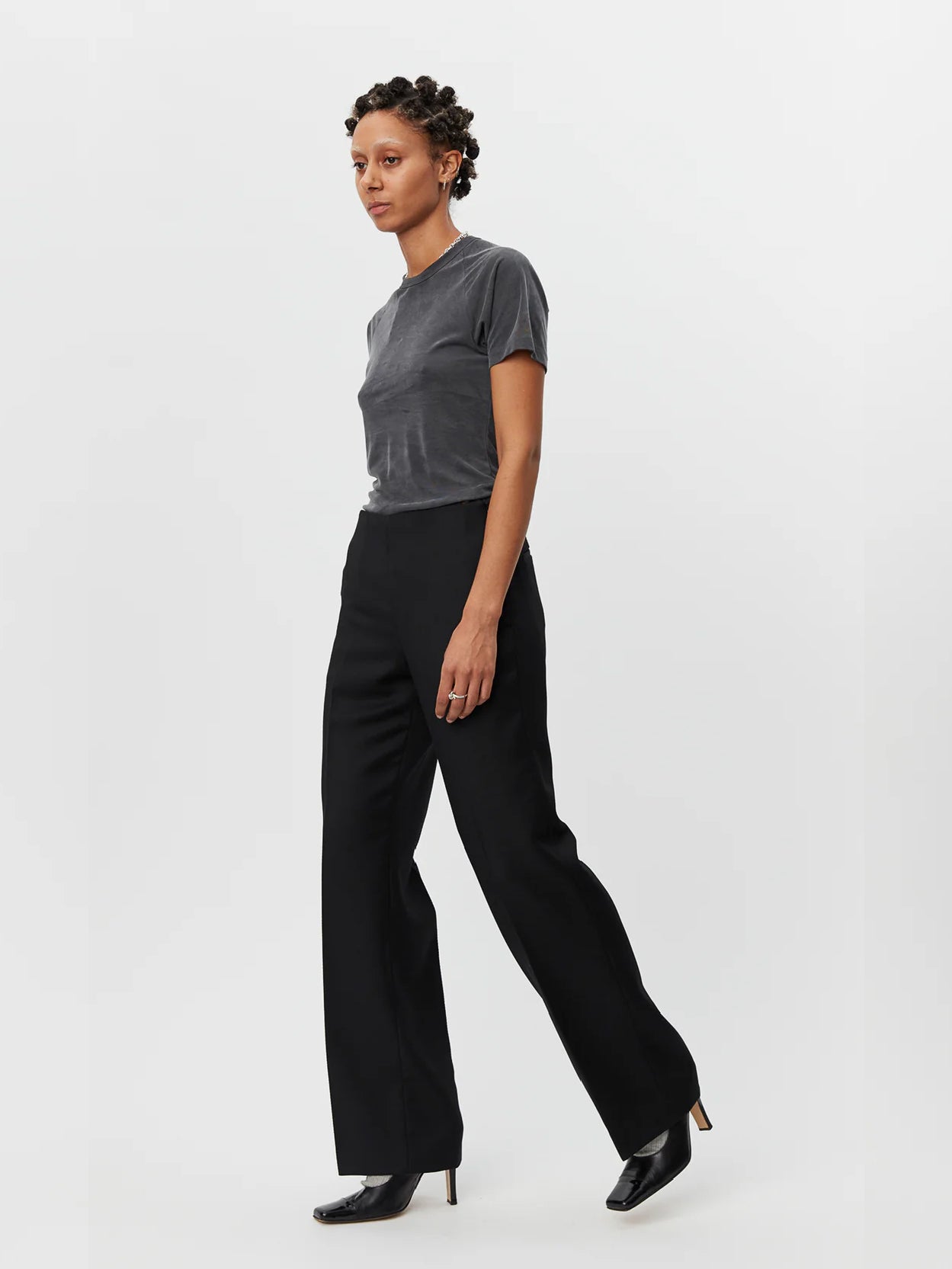 A person stands against a plain white background, wearing a fitted gray short-sleeve T-shirt and black Taper Trousers Black by Mfpen made from virgin wool. They have short hair styled in twists and are wearing black shoes with a small heel.