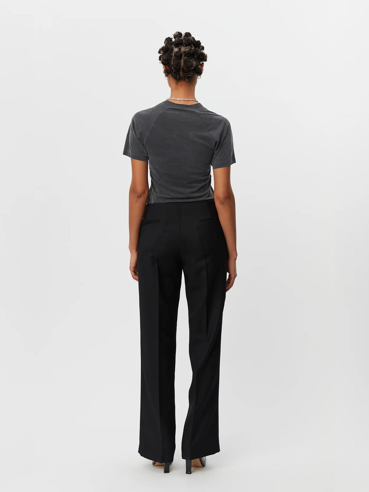 A person stands against a plain white background, wearing a fitted gray short-sleeve T-shirt and black Taper Trousers Black by Mfpen made from virgin wool. They have short hair styled in twists and are wearing black shoes with a small heel.