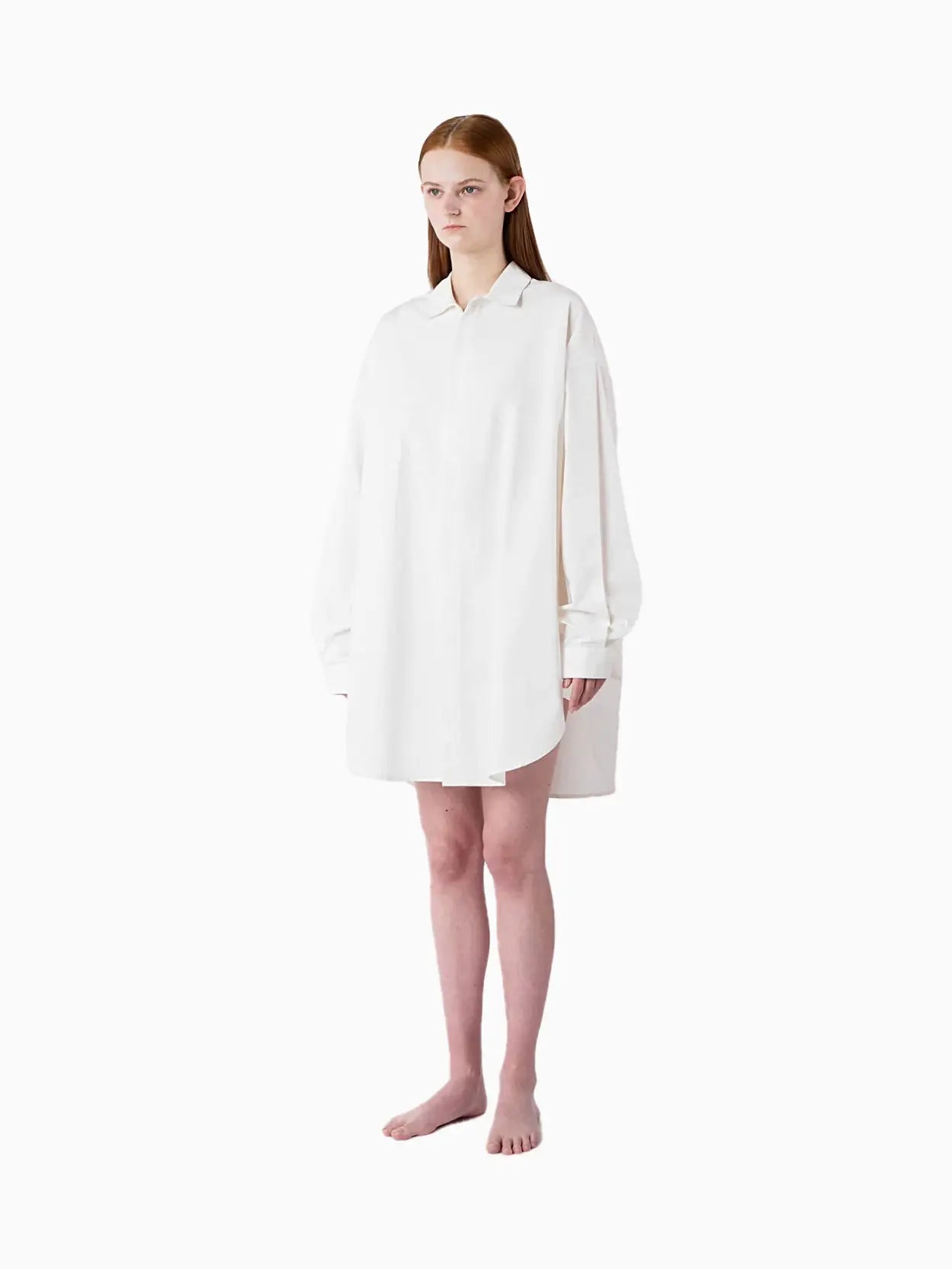 A white button-up shirt with long sleeves, a collar, and a loose, slightly oversized fit. The Mega Overshirt White has a minimalist design and appears to be made from lightweight fabric. A small tag with text is visible at the back of the collar, subtly nodding to its origins from Sunnei in Barcelona.