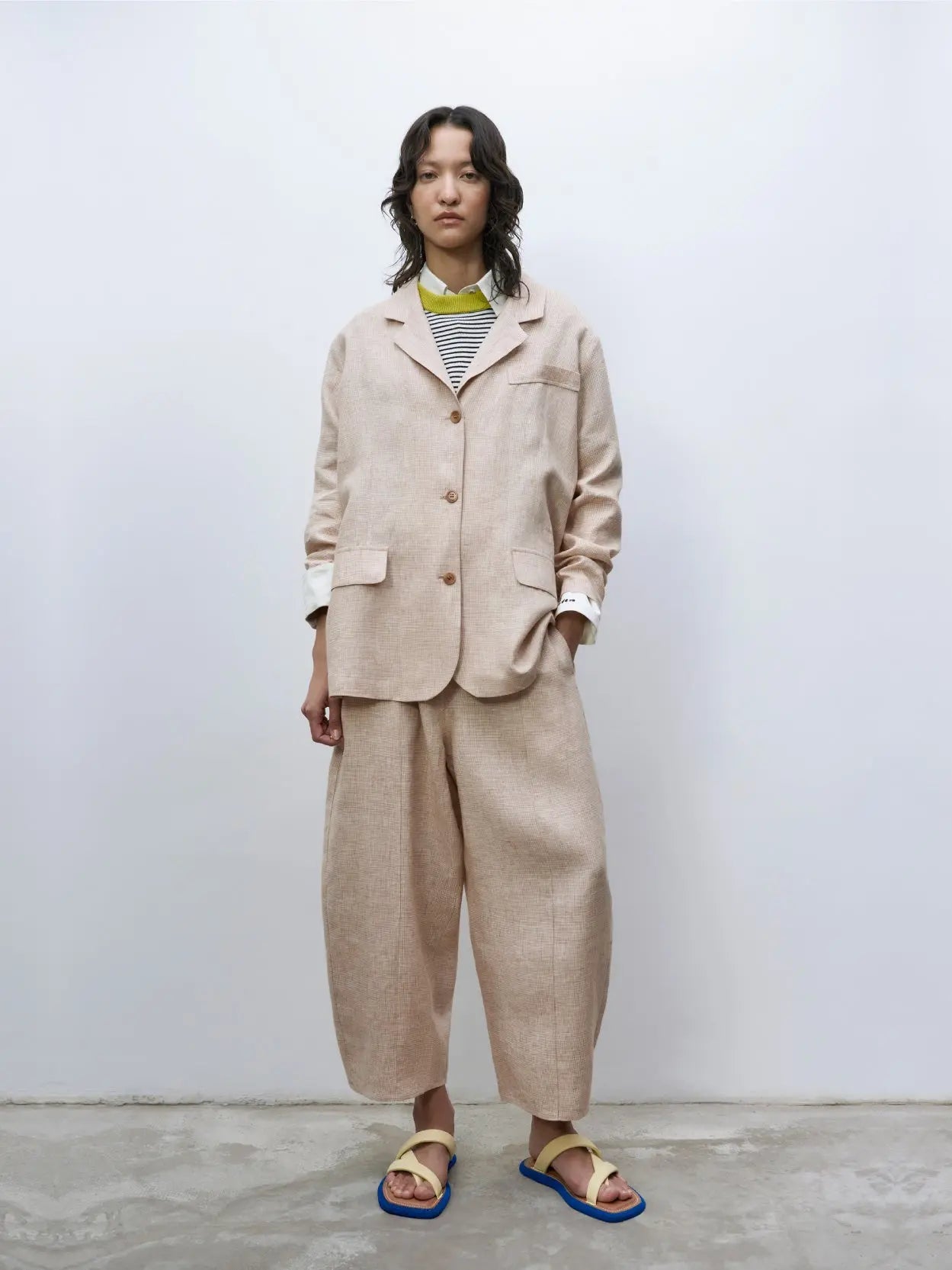 A chic pair of beige, loose-fitting Melange Linen Curved Pants from Cordera in Barcelona, featuring an elastic waistband and a button detail on the front. The straight-cut design has vertical seam lines running down each leg and ends just above the ankles, offering a stylish and comfortable fit.