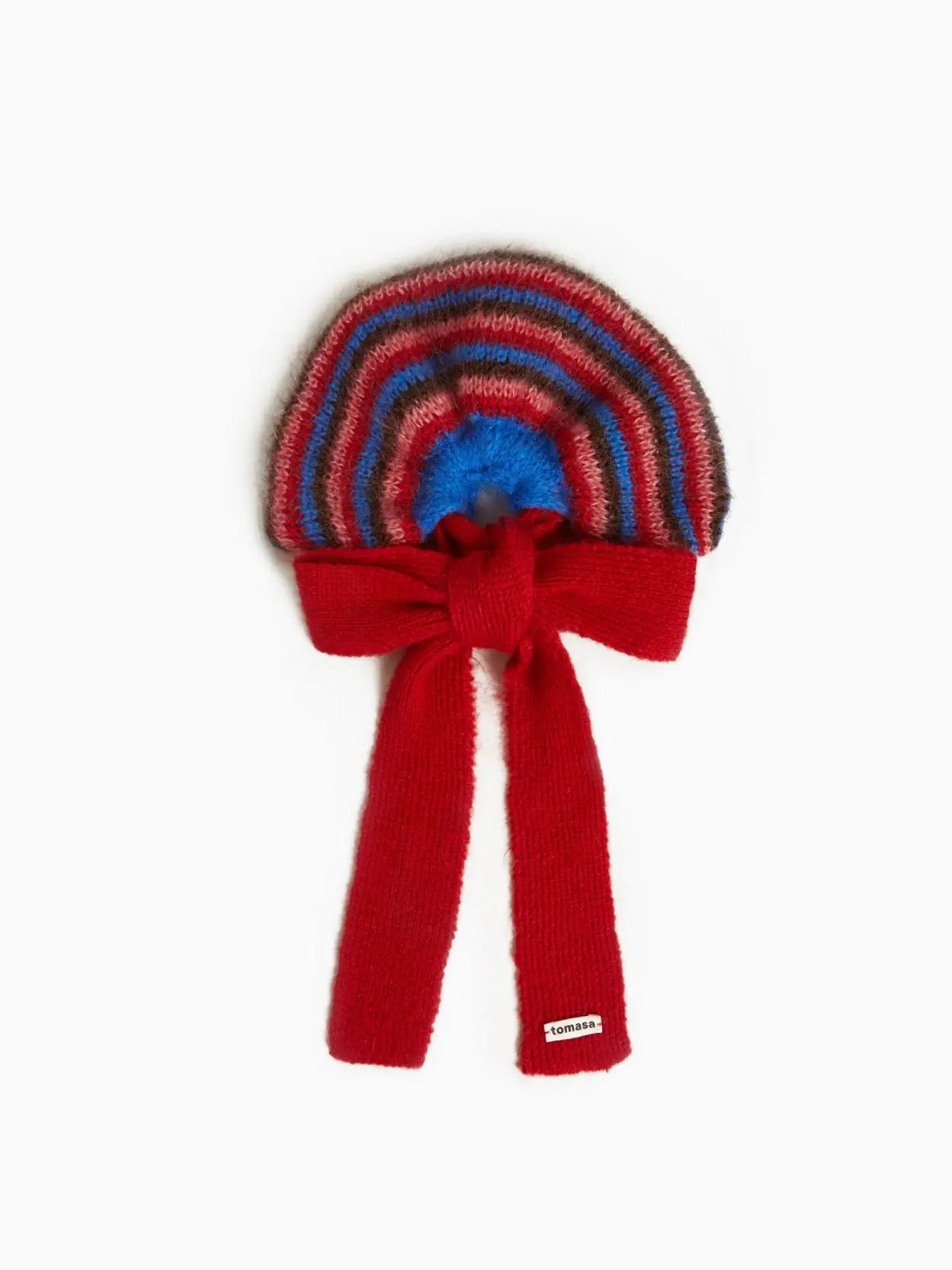 A colorful striped Mencia Bow Scrunchie with a knitted red bow at the base and a red fringe. The stripes include shades of red, blue, and brown, forming a circular pattern. A small white label with the word "Tomasa" is attached at the bottom of the bow. Find it exclusively at Bassalstore in Barcelona.
