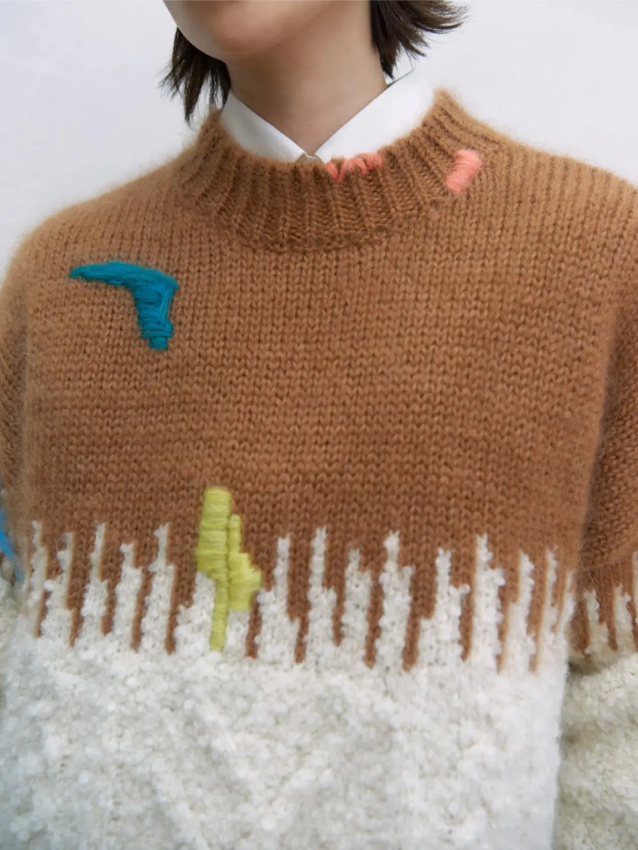 A person with short hair is wearing a cozy Cordera Mohair Embroidered Sweater with a brown top portion, white lower portion featuring a textured pattern, and colorful abstract designs. They are standing against a plain background, looking directly at the camera, perhaps right outside bassalstore in Barcelona.