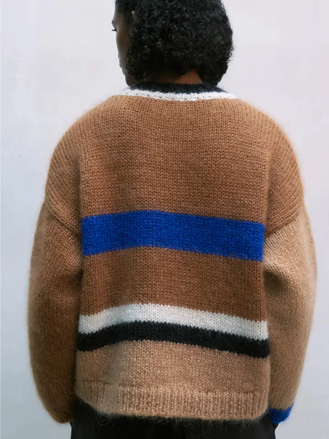 A Mohair Striped Sweater from Cordera featuring a brown base with wide horizontal stripes in blue, white, and black across the chest. The long sleeves and body have a loose, boxy fit, and there is black trim around the neckline. The garment is laid flat against a white background, ready for your wardrobe in Barcelona.