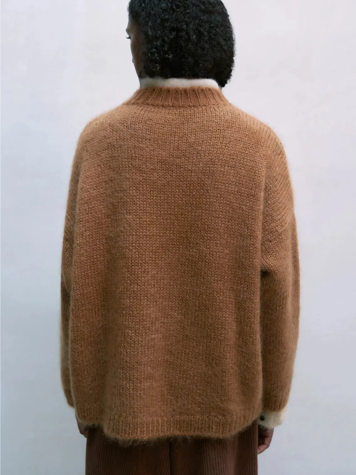 A person with shoulder-length curly hair is wearing a loose-fitting Mohair Sweater Toffee by Cordera over a white collared shirt and brown corduroy pants. They have their hands in their pockets and are standing against a plain light-colored background, looking as if they just stepped out of a stylish boutique in Barcelona.