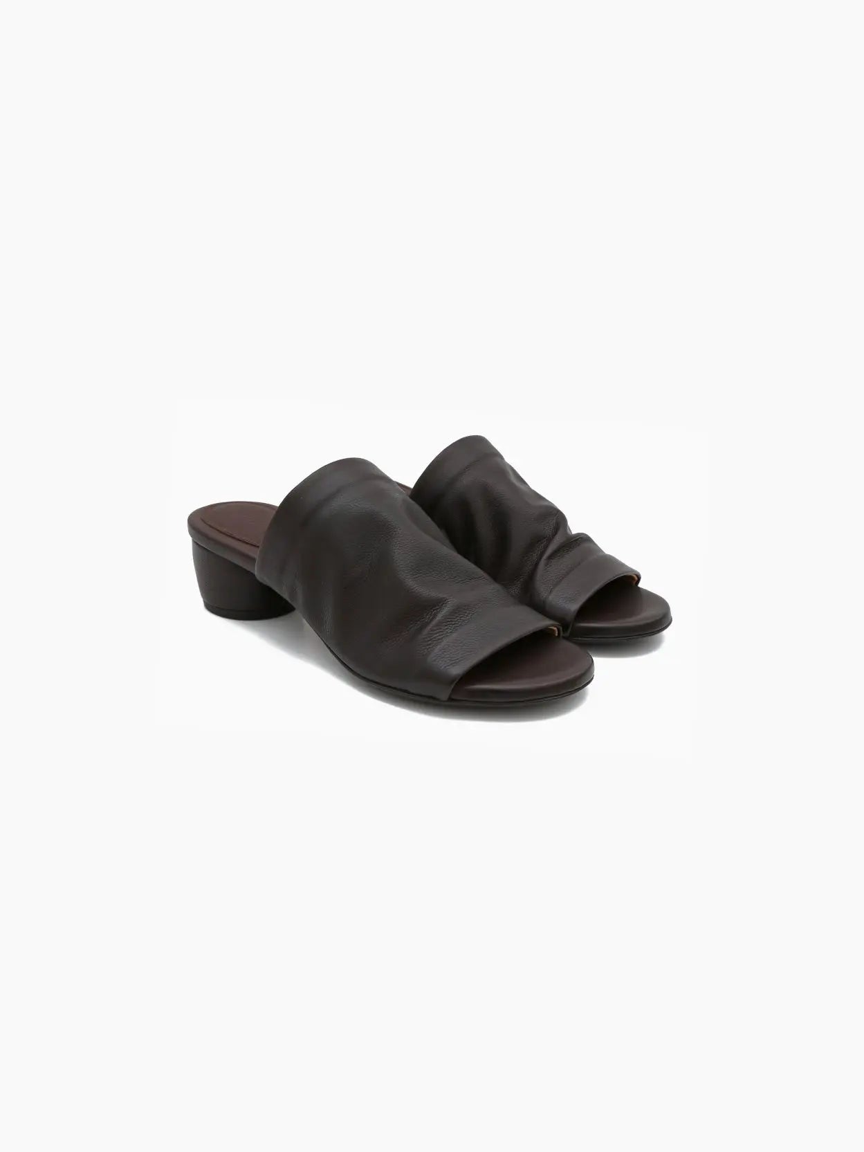 A dark brown mule-style sandal with a wide strap covering the upper foot and an open toe, available at Bassalstore. The Otto Sandal Dark Brown features a low, chunky heel and a minimalist design. Pictured against a white background in a side view, this shoe embodies chic Barcelona fashion from Marsèll.