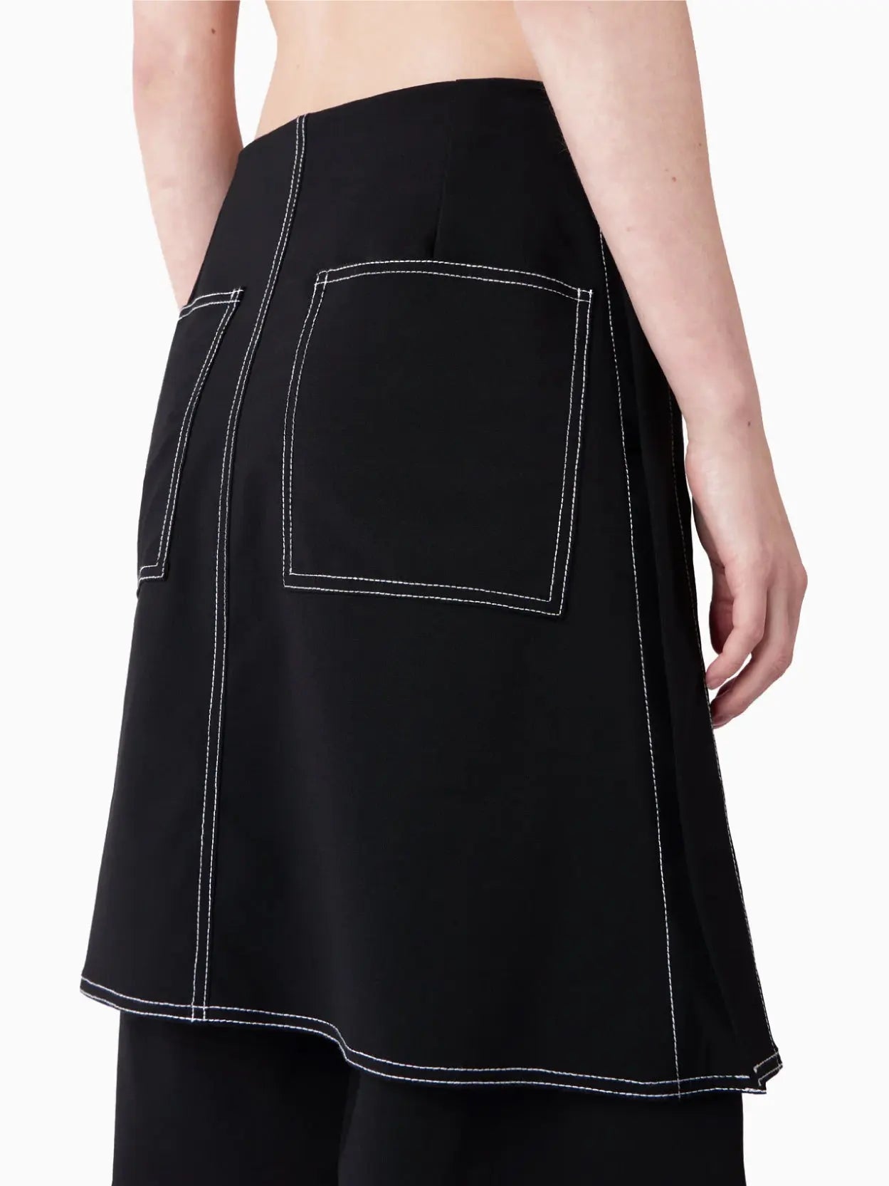 A black double-layered mini skirt with white stitching accents. The design features a layered look, with a shorter top layer and a longer underlayer, creating a stylish and modern appearance. Displayed against a white background, this chic piece is available at Bassalstore in Barcelona. The product name is Panta Skirt Black by Sunnei.