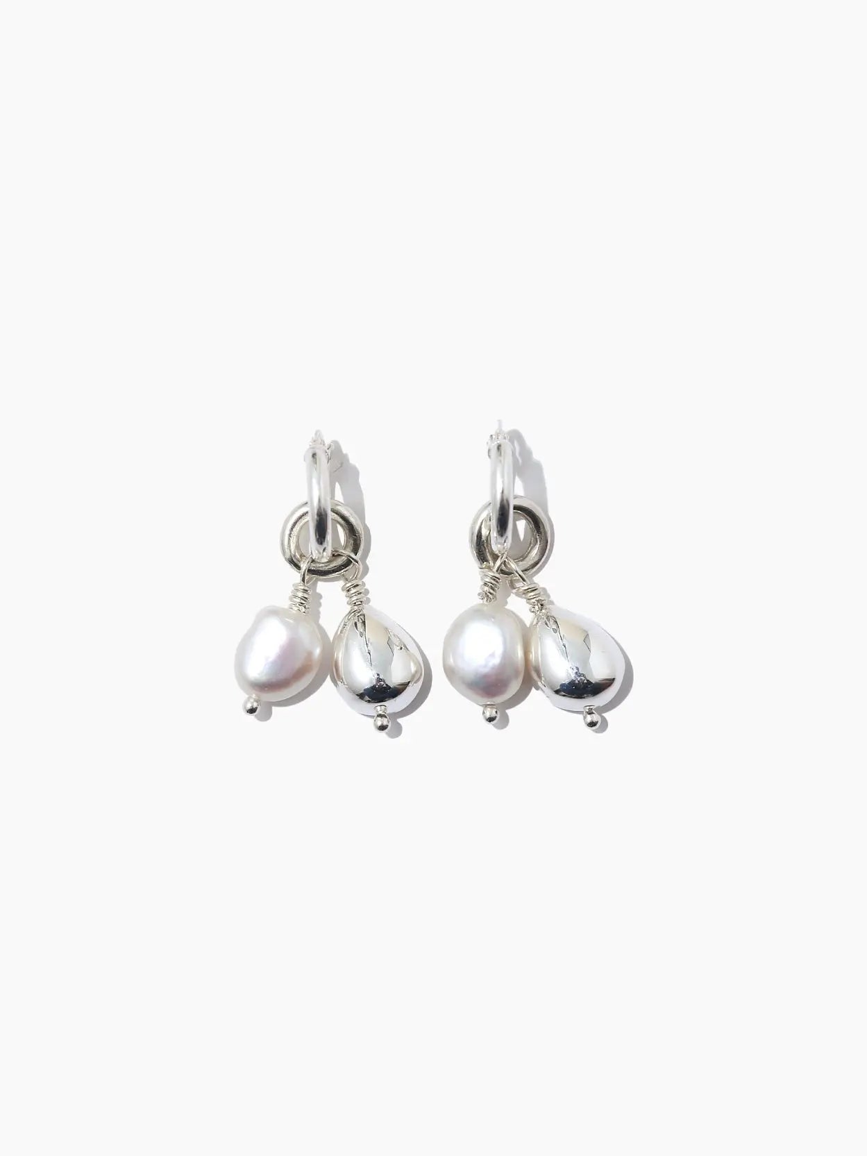 A pair of Perla Hoops by Nathalie Schreckenberg with hanging irregularly shaped white pearls. The pearls are attached to the hoops by small silver loops, creating a delicate and elegant design. Set against a plain white background, these earrings available at Bassalstore in Barcelona highlight simplicity and beauty.