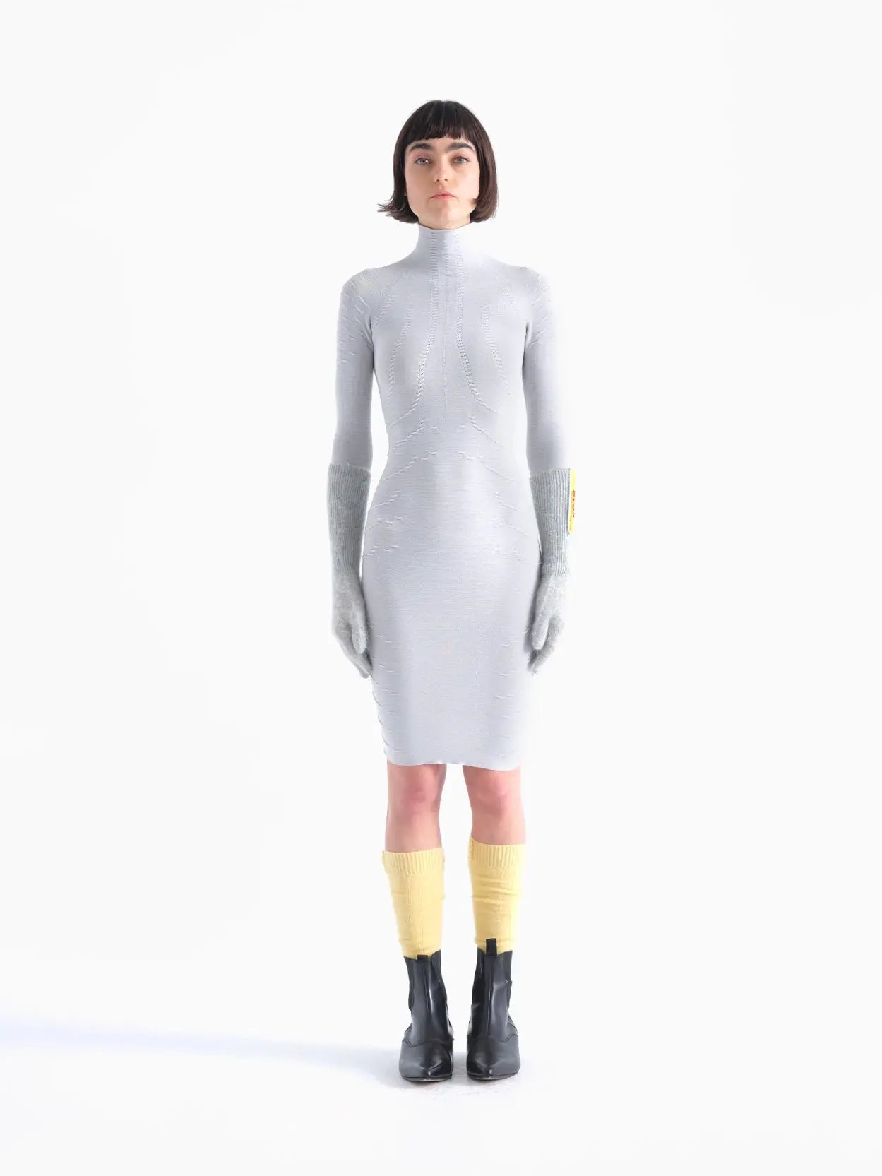A person with short hair stands facing forward on a plain white background. They are dressed in a form-fitting, long-sleeved, light gray Rai Dress Grey from Bielo, with large gray mittens covering their hands, yellow leg warmers, and black ankle boots.