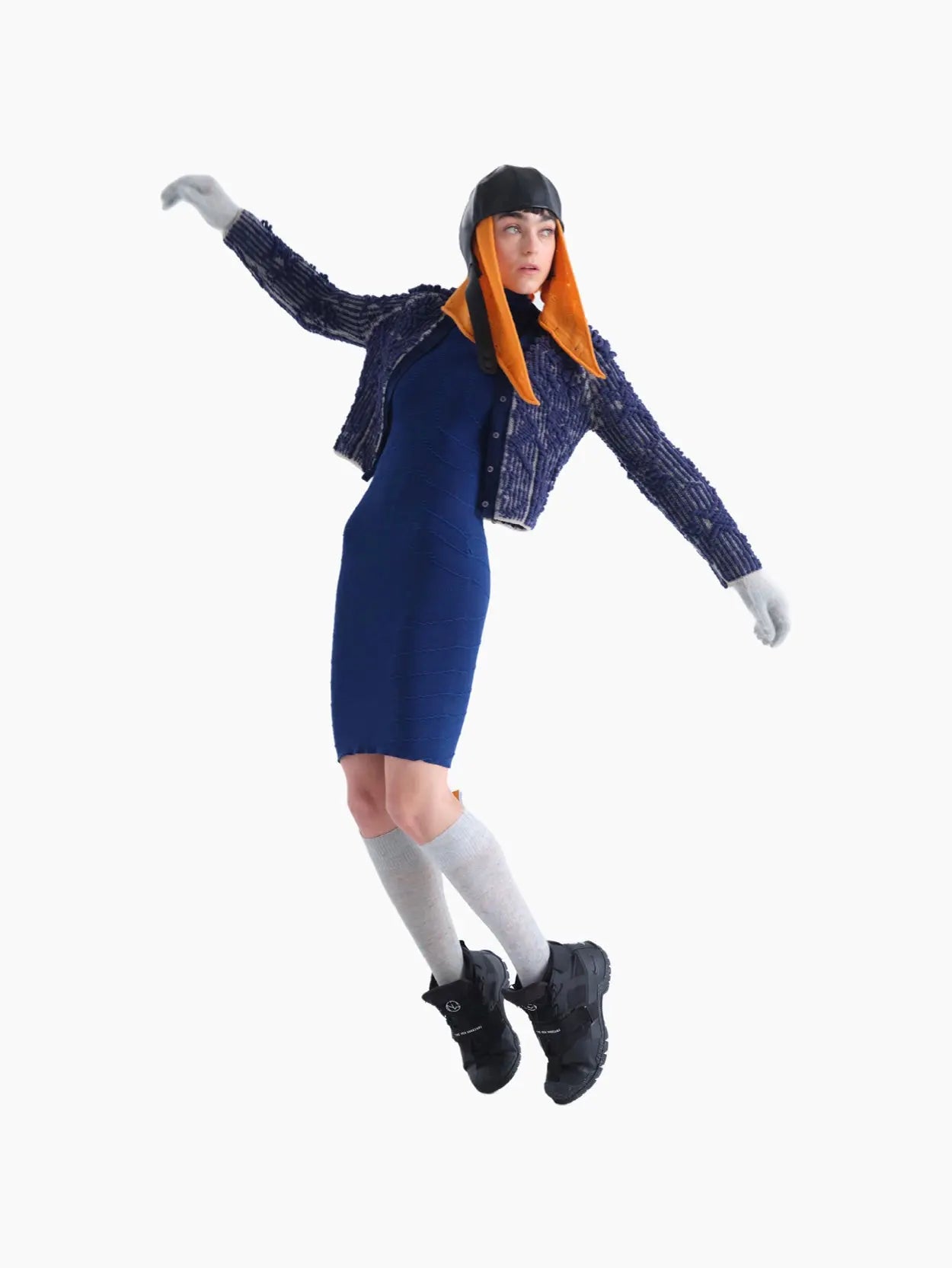 A person is standing against a white background wearing a blue textured jacket, a Rai Dress Navy by Bielo, gray gloves, gray knee-high socks, black boots, and a unique hat with orange ear flaps that cover the sides of their face. They are facing forward with a neutral expression, looking like they just stepped out of an exclusive Barcelona store.