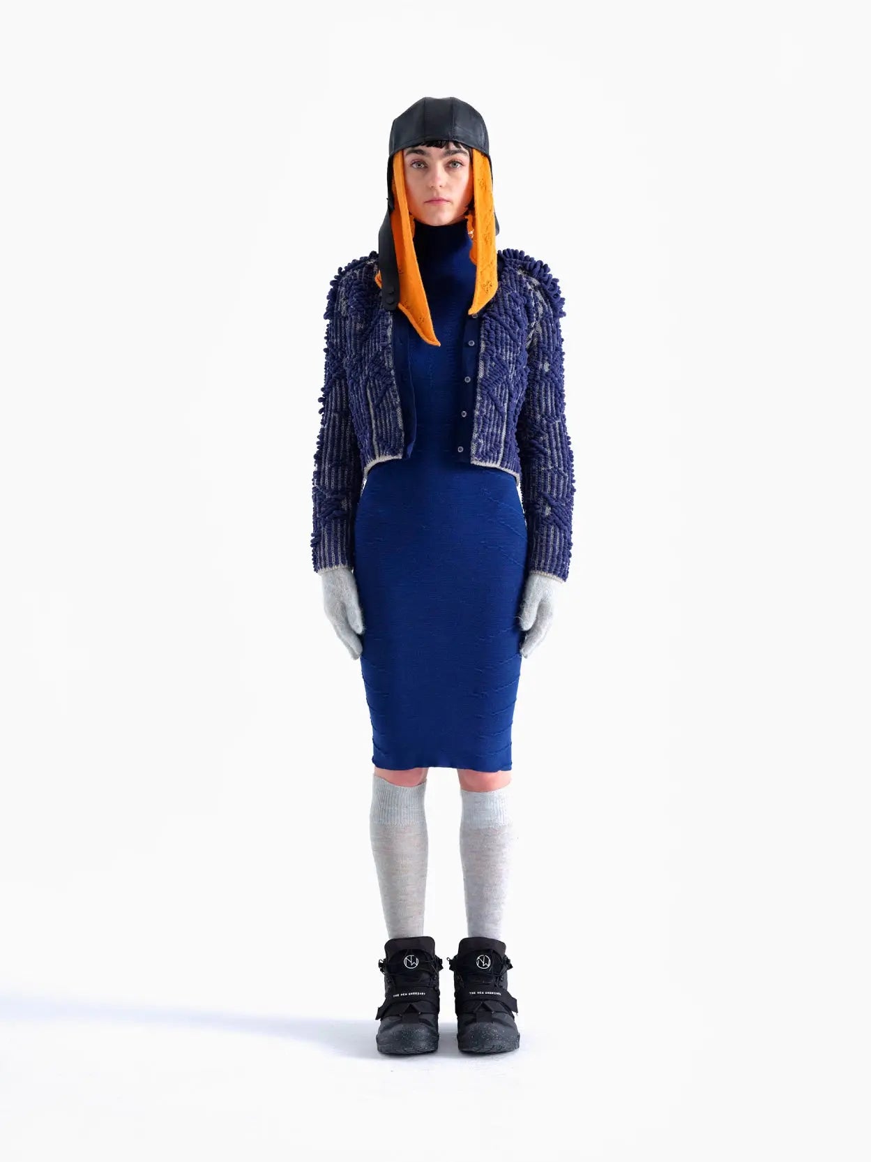 A person is standing against a white background wearing a blue textured jacket, a Rai Dress Navy by Bielo, gray gloves, gray knee-high socks, black boots, and a unique hat with orange ear flaps that cover the sides of their face. They are facing forward with a neutral expression, looking like they just stepped out of an exclusive Barcelona store.