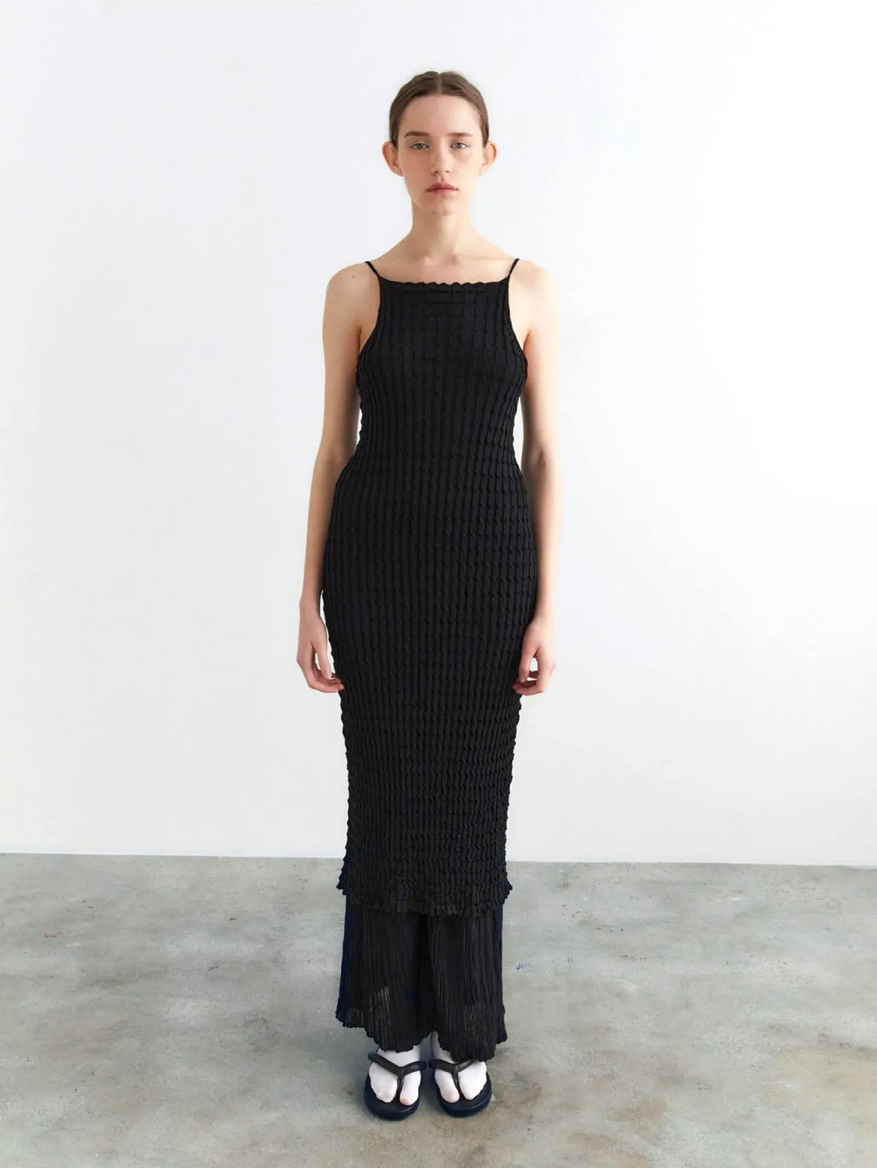 A long, sleeveless black dress with thin shoulder straps is displayed against a plain white background. The Rina Dress Black by Rus, featured at Bassalstore in Barcelona, boasts textured, crinkled fabric and a straight, narrow silhouette.