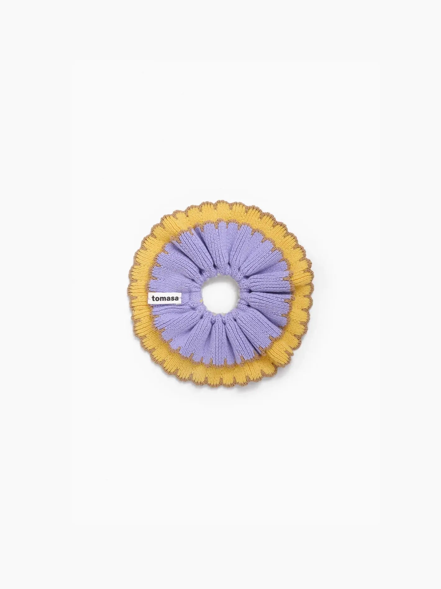 A circular, two-tone scrunchie called the "Ruffle Scrunchie Amanecer" by Tomasa is displayed against a white background. The outer part is yellow, and the inner part is purple with a ridged design. A small fabric label with the text "Tomasa" is attached to the inner part of the scrunchie, available exclusively at Bassalstore in Barcelona.