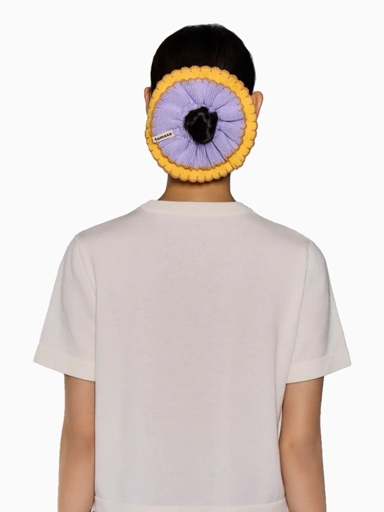 A circular, two-tone scrunchie called the "Ruffle Scrunchie Amanecer" by Tomasa is displayed against a white background. The outer part is yellow, and the inner part is purple with a ridged design. A small fabric label with the text "Tomasa" is attached to the inner part of the scrunchie, available exclusively at Bassalstore in Barcelona.