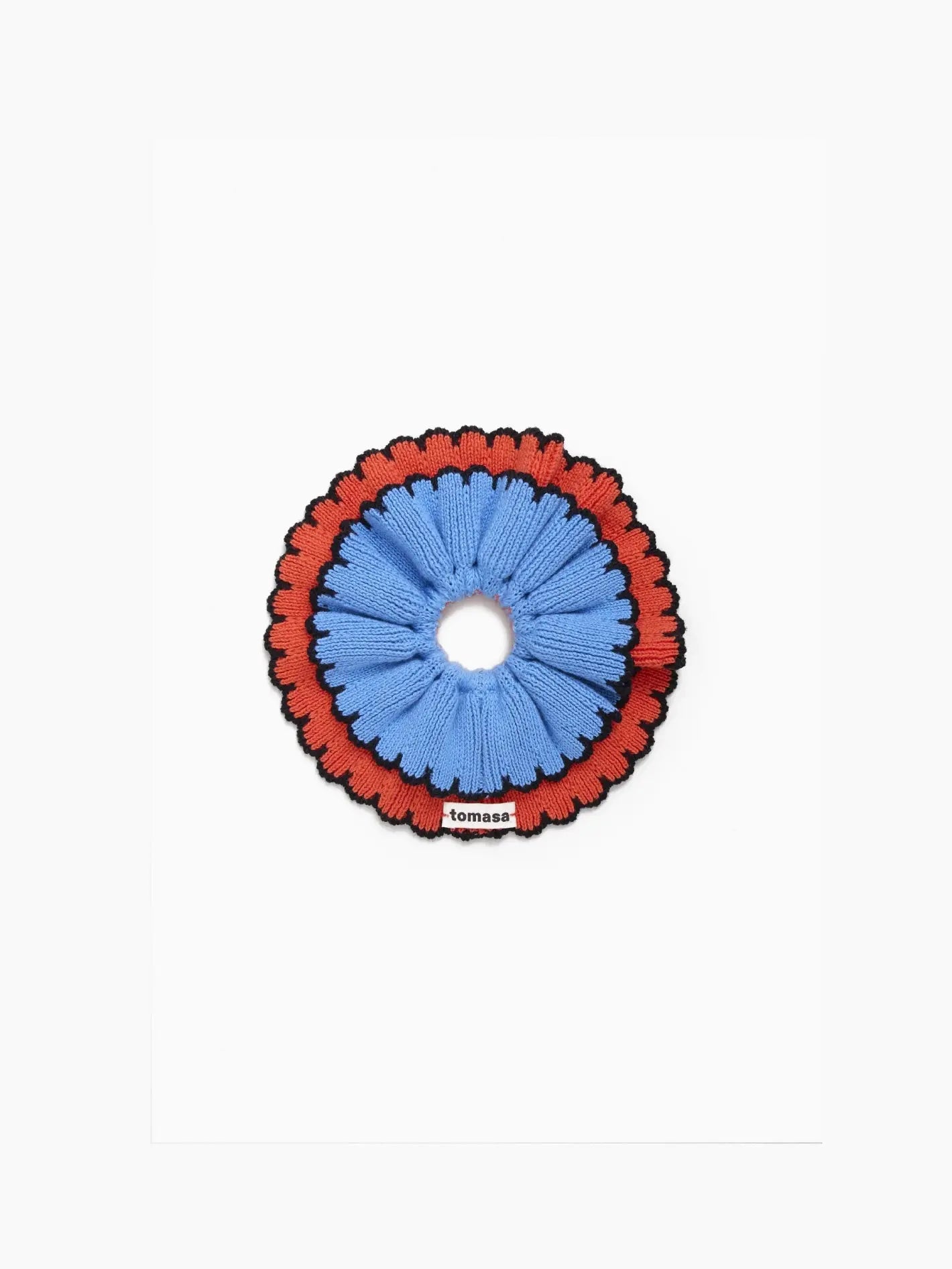 A round, fabric hair scrunchie in blue and red colors. The inner portion of the scrunchie is blue, while the outer ring is red with a decorative edge. A small label with the text "Tomasa" is visible at the bottom. The Ruffle Scrunchie Lumbre, available at Bassalstore in Barcelona, rests on a plain white background.