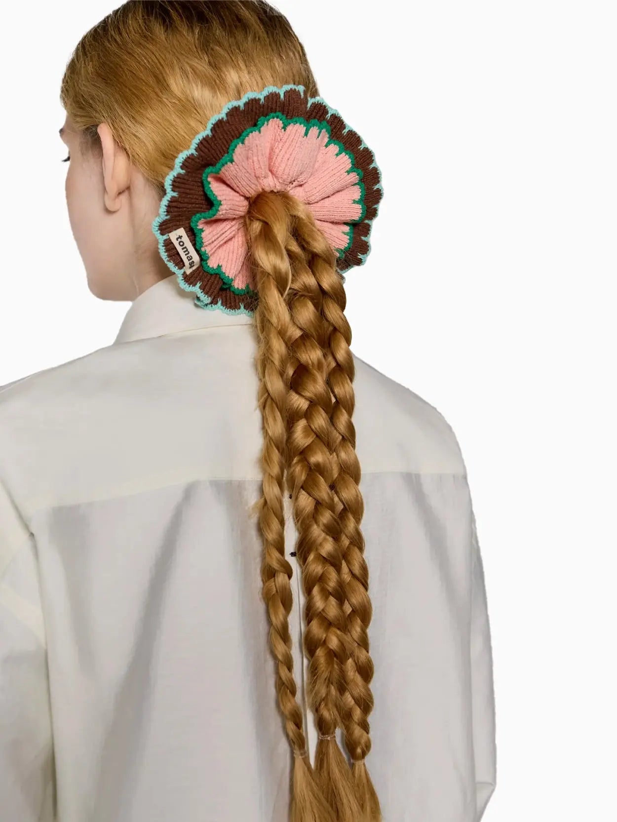A Ruffle Scrunchie Tierra with a soft pink center, edged with dark brown and a thin green trim. A small white tag labeled "Tomasa" is attached to the scrunchie, available at Bassalstore in Barcelona. The background is plain white.