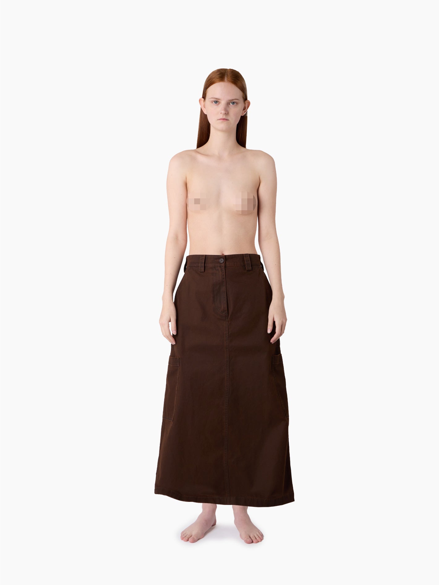 A person with long hair stands barefoot against a plain background, wearing only a Sunnei Cargo Skirt Brown made in Italy. Their upper body is bare with nipples censored.