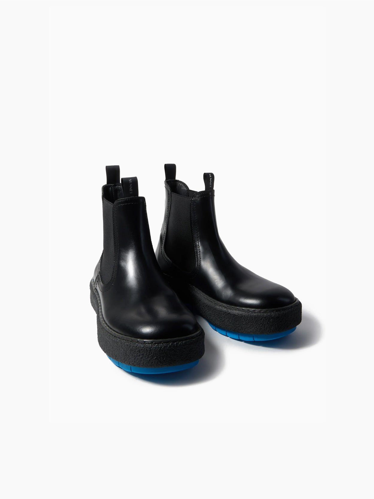 The Cemento Chelsea Boot by Sunnei is a black leather ankle boot with a chunky textured sole. It features black detailing and a vibrant blue tread, along with elastic side panels and pull tabs for easy wear, all set against a plain white background.