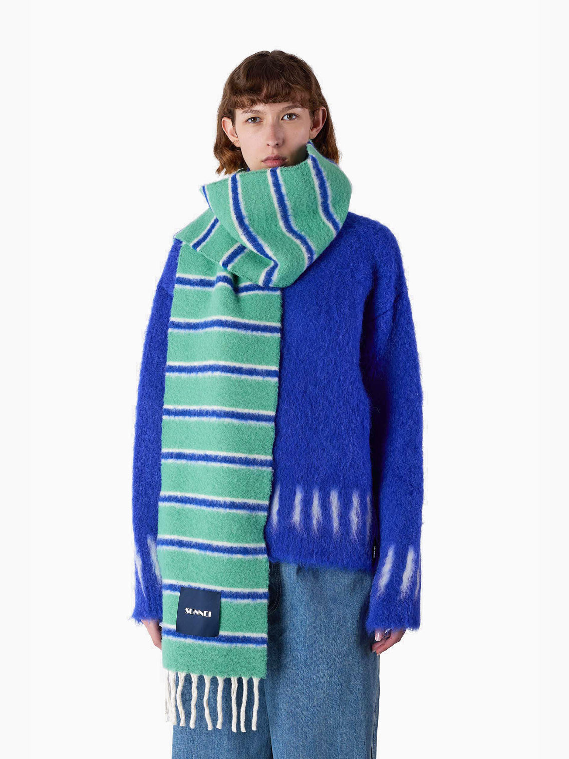 The Green Stripes Scarf by Sunnei, elegantly folded, is crafted from luxurious alpaca wool and features blue and white stripes against a green background. It rests gracefully on a white backdrop, boasting a black label with white text and finished with fringes at the end. Proudly made in Italy.