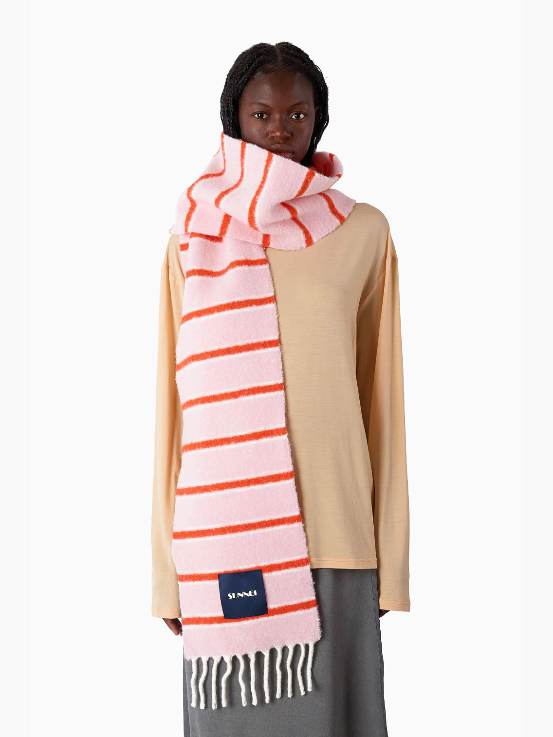 The Pink Stripes Scarf by Sunnei, made from luxurious alpaca wool, is artistically designed with pink and red stripes and accented with white fringes at the edges. Elegantly folded, it bears a dark blue label with white text. Crafted in Italy, this exquisite piece stands out beautifully against a plain white background.