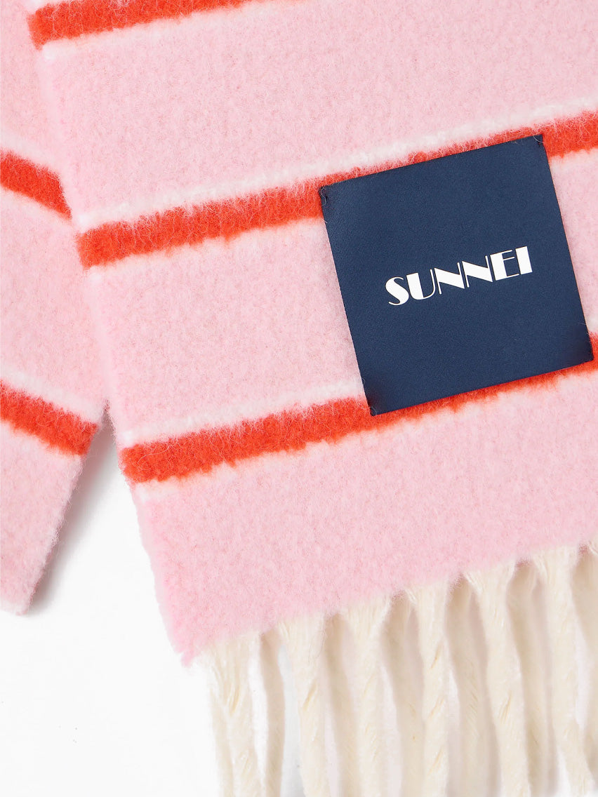 The Pink Stripes Scarf by Sunnei, made from luxurious alpaca wool, is artistically designed with pink and red stripes and accented with white fringes at the edges. Elegantly folded, it bears a dark blue label with white text. Crafted in Italy, this exquisite piece stands out beautifully against a plain white background.