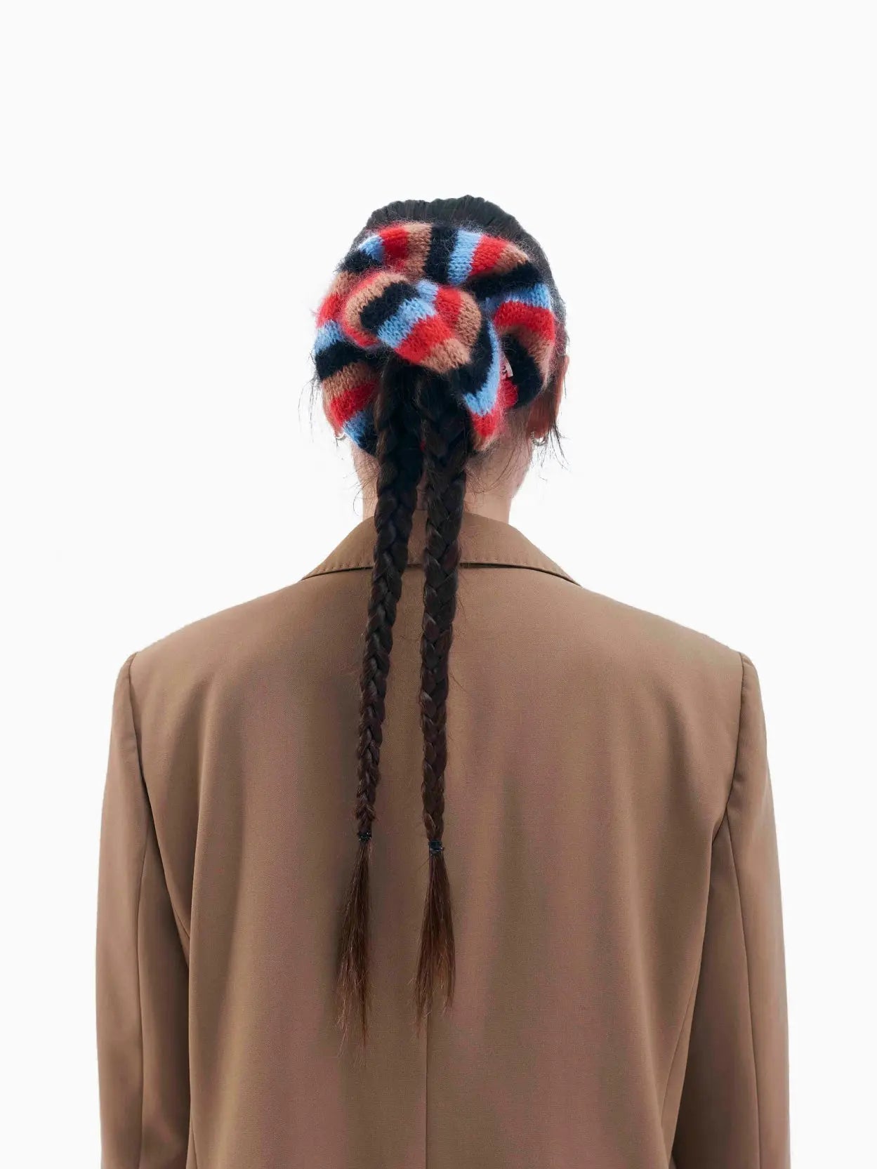 A round, striped wool or knitted hair scrunchie with alternating red, blue, black, and tan colors. The Serena Scrunchie has a small brand tag from Tomasa attached to it. Photographed against a plain white background, it's a vibrant accessory that captures the essence of Barcelona style.