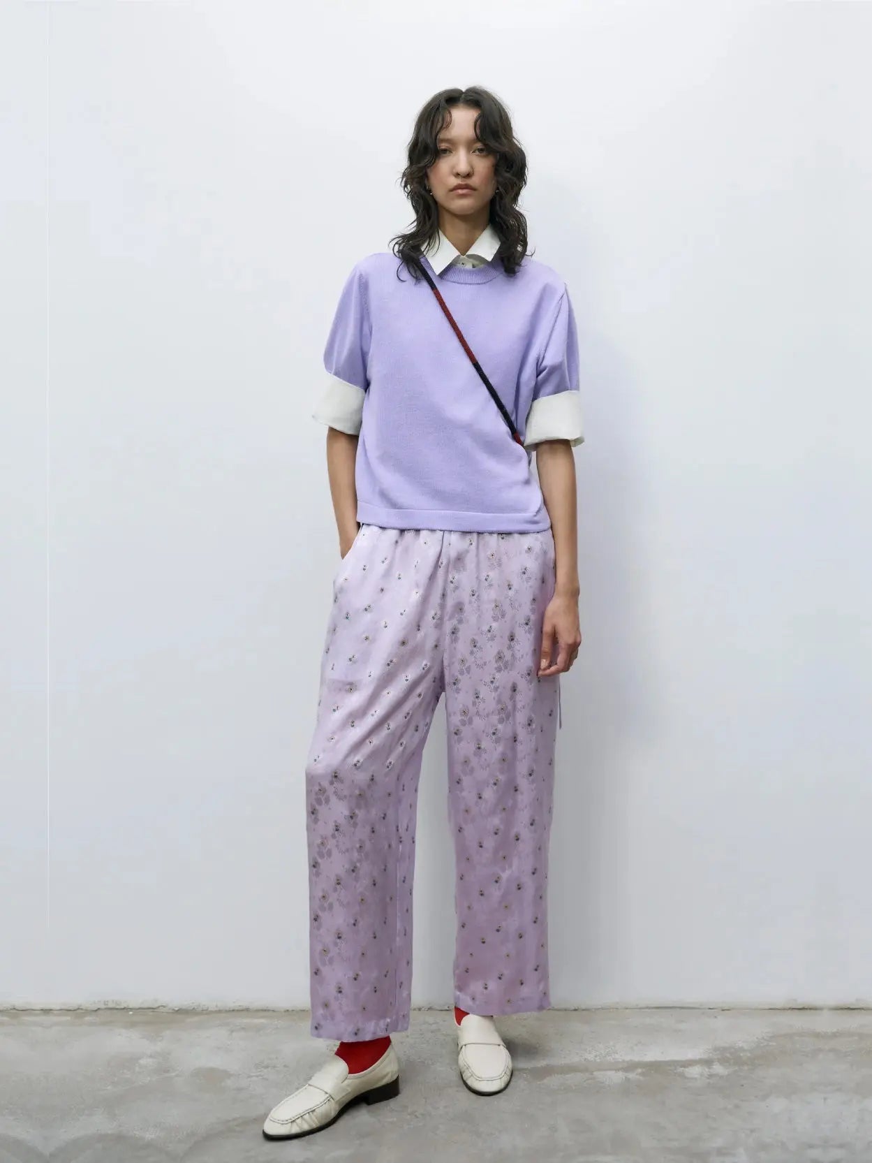 A person stands against a plain white background. They are dressed in a light purple, short-sleeve sweater over a white shirt, Silk Floral Pants Cardo from Cordera, white shoes, and red socks. A small crossbody bag from bassalstore in Barcelona is slung over their shoulder.