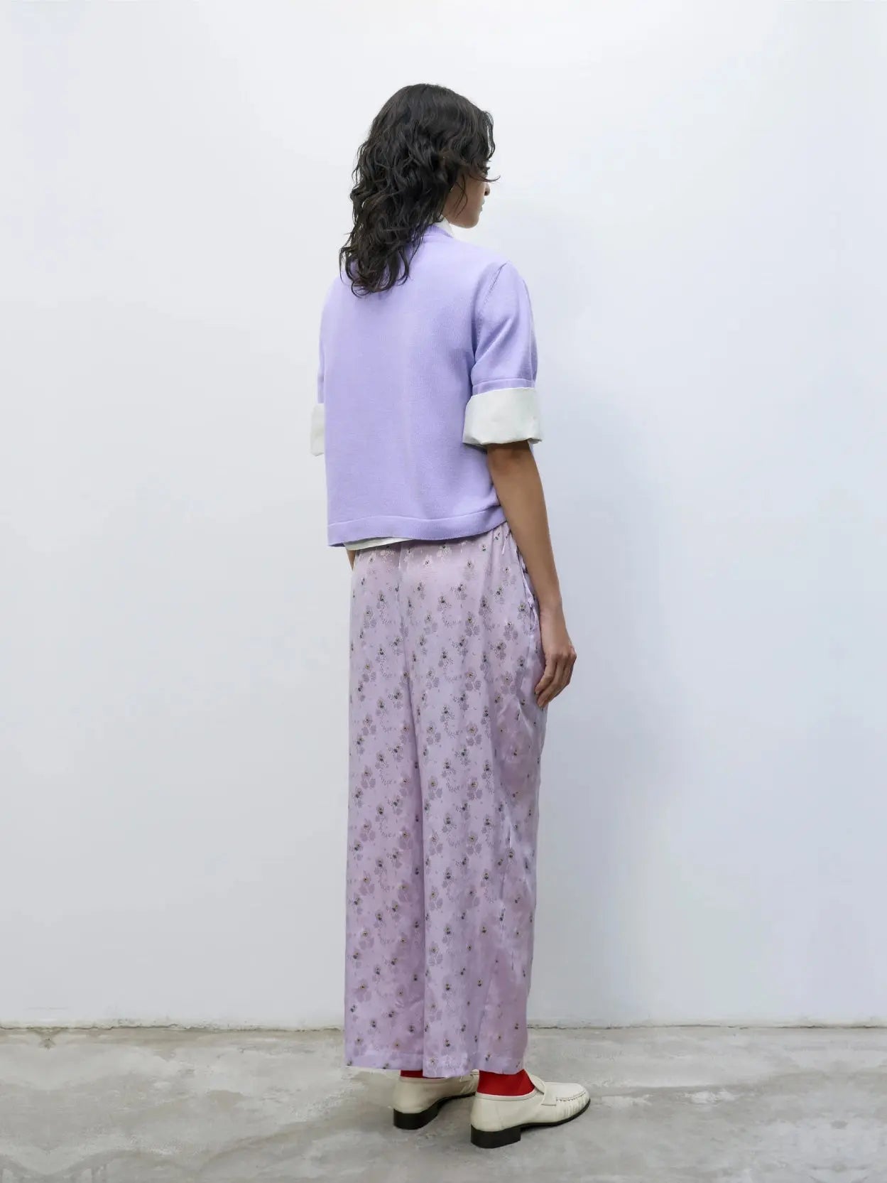 A person stands against a plain white background. They are dressed in a light purple, short-sleeve sweater over a white shirt, Silk Floral Pants Cardo from Cordera, white shoes, and red socks. A small crossbody bag from bassalstore in Barcelona is slung over their shoulder.