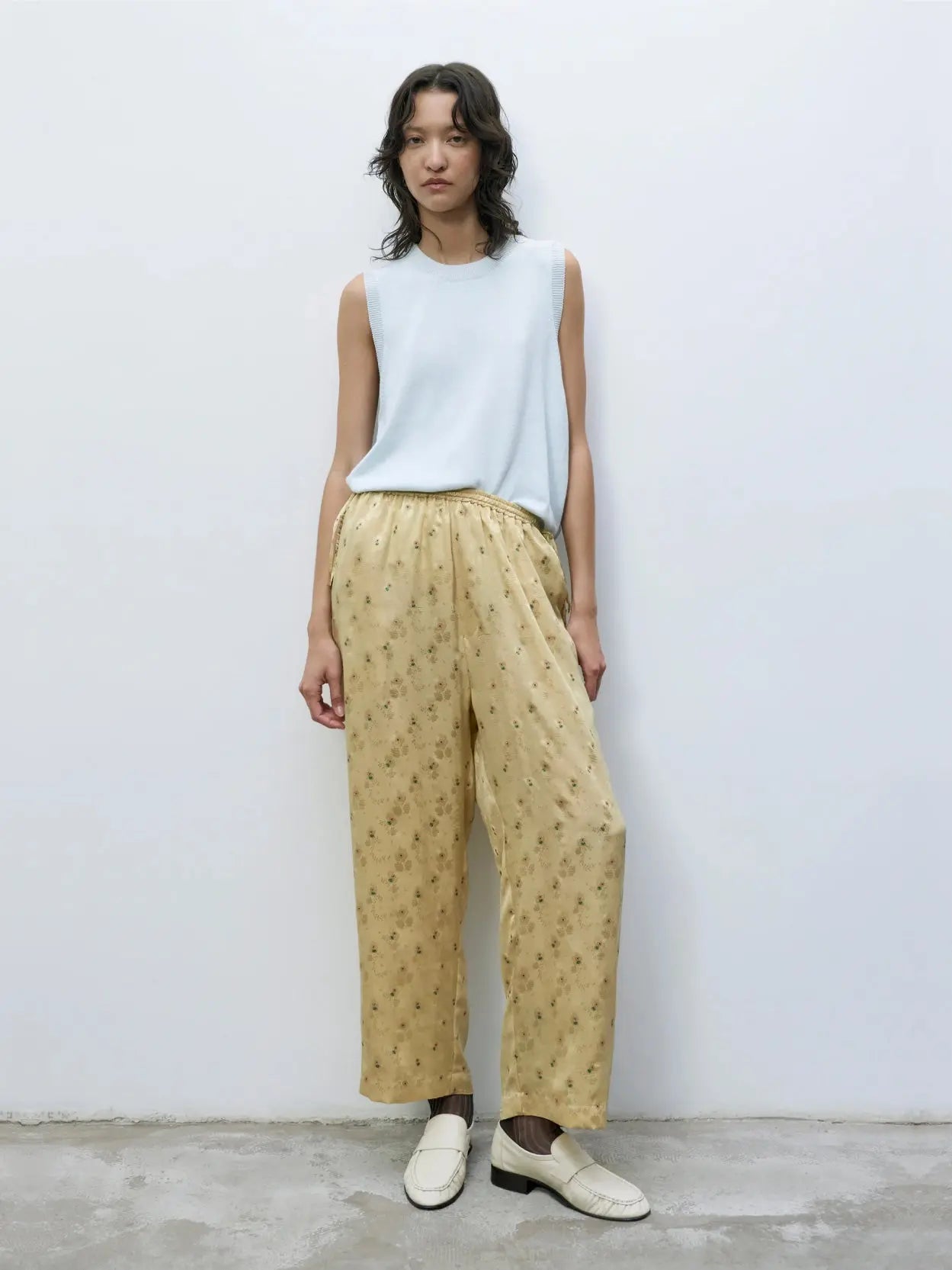 A person with shoulder-length wavy hair stands against a plain white wall in front of the Bassal store in Barcelona. They are wearing a sleeveless light blue top and Cordera's Silk Floral Pants Jojoba. They also have on white loafers. The flooring is gray.