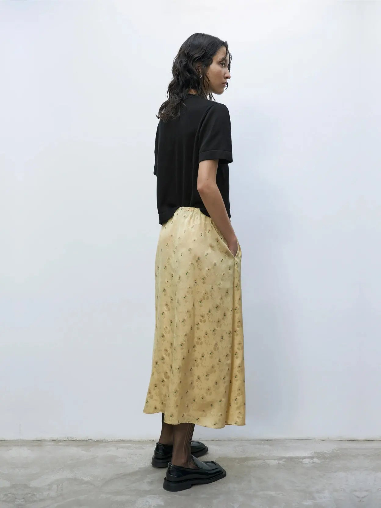 A woman with long wavy hair stands against a plain white wall. She is wearing a loose-fitting black T-shirt from BassalStore, a Silk Floral Skirt Jojoba from Cordera, and black loafers. Her expression is neutral, and her hands are relaxed at her sides.