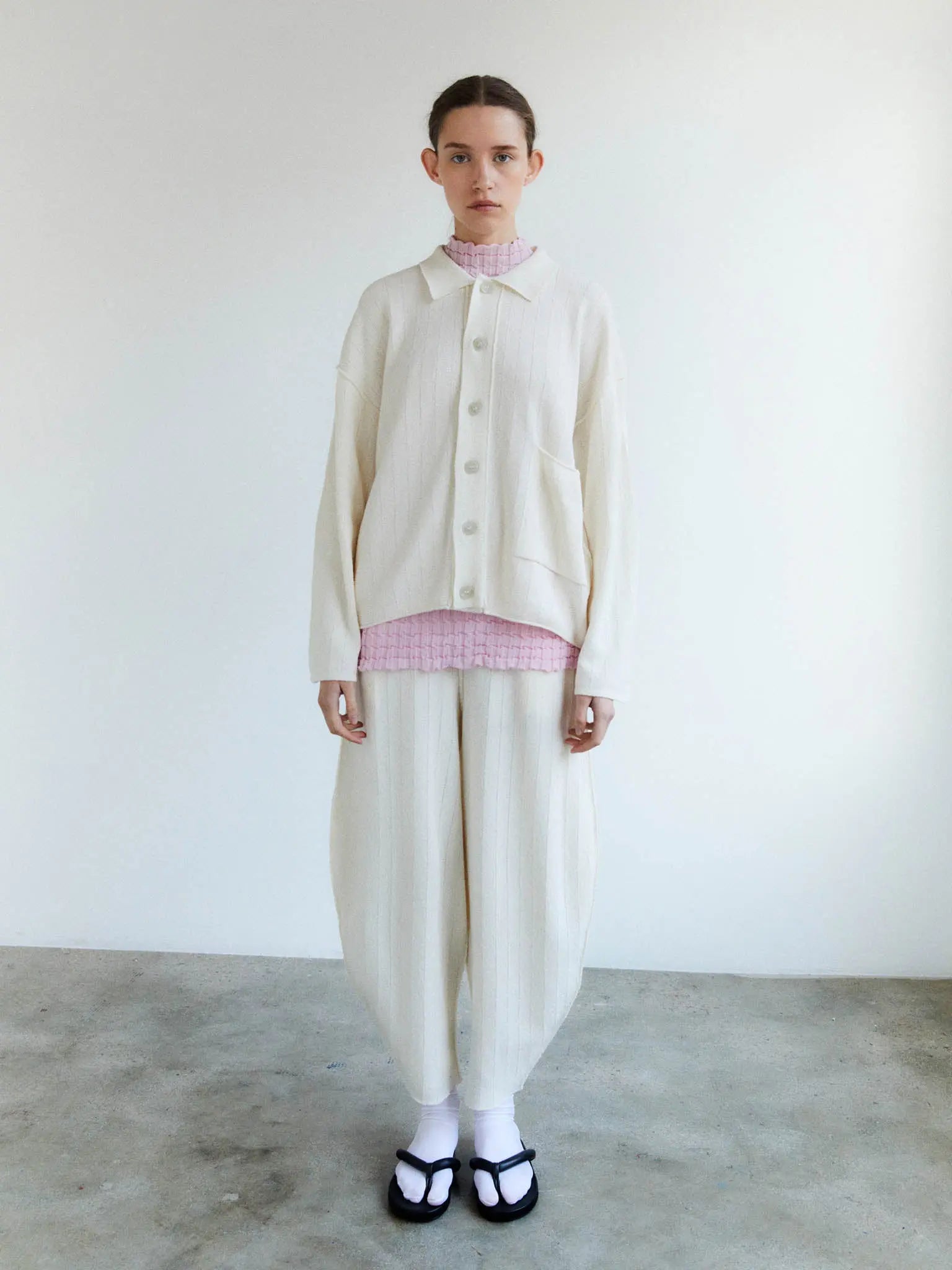 A cream-colored, long-sleeve button-up cardigan with a ribbed texture. It features a collar, front buttons, and a single large pocket on the left side. The label inside the neckline reads "Rus." Laid flat against a plain white background, this stylish piece is available at Bassalstore in Barcelona. This is the Tobira Cardigan Chalk by Rus.