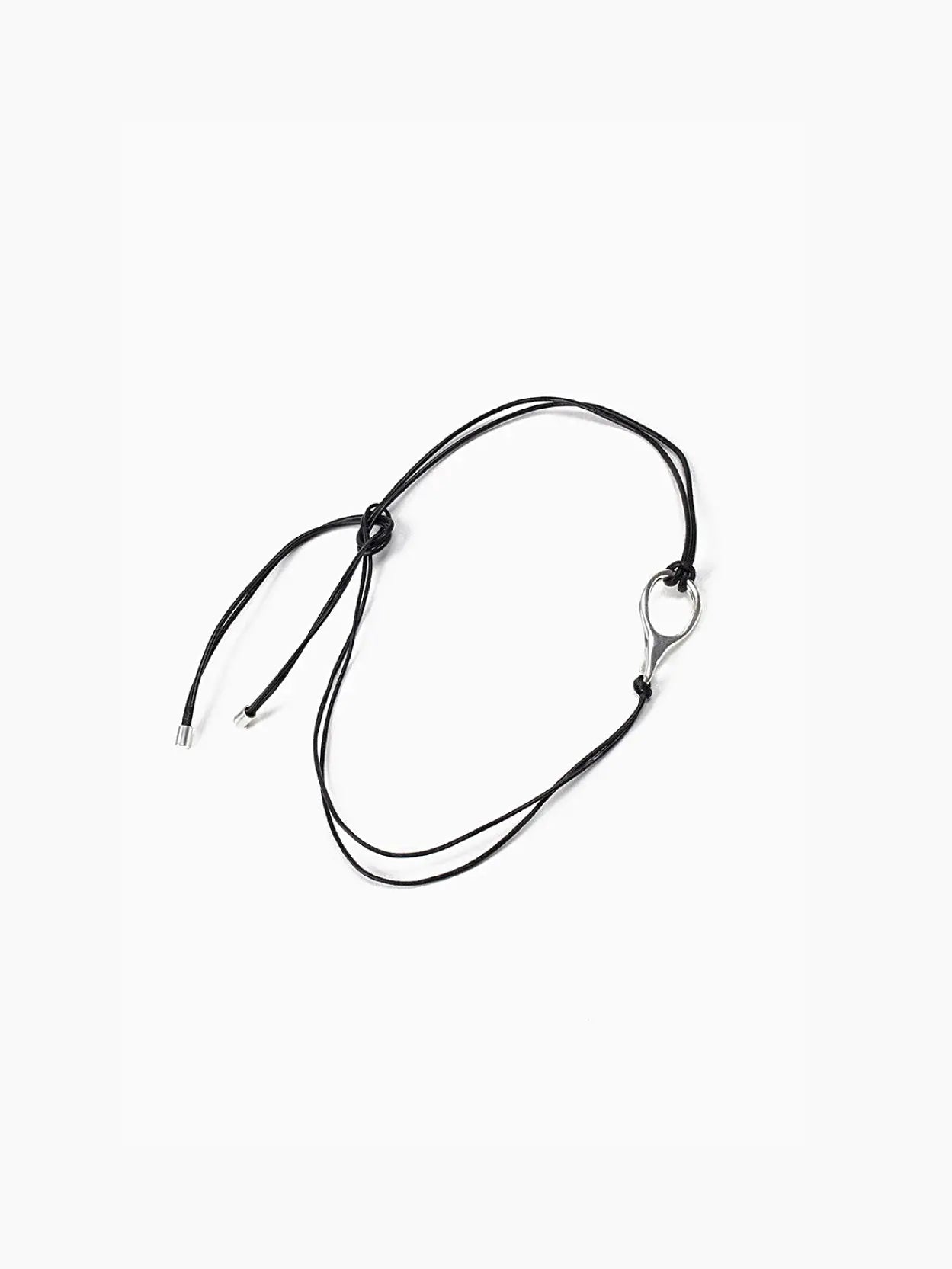A black, minimalist Toggle Necklace placed on a white background. The necklace, designed by Nathalie Schreckenberg and available at Bassalstore in Barcelona, features adjustable knots and has a small, silver oval charm looped through the cord. The ends of the cord have metallic tips.