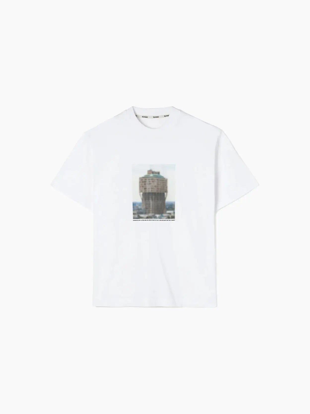 A Torre Velasca T-Shirt Re-Edition available at Bassalstore, featuring a central graphic image of a tall, cylindrical building situated by the water. The architectural structure is prominently displayed on the front of the shirt against the solid white background, capturing a Barcelona-inspired vibe. This stylish piece from Sunnei perfectly merges iconic architecture with contemporary fashion.