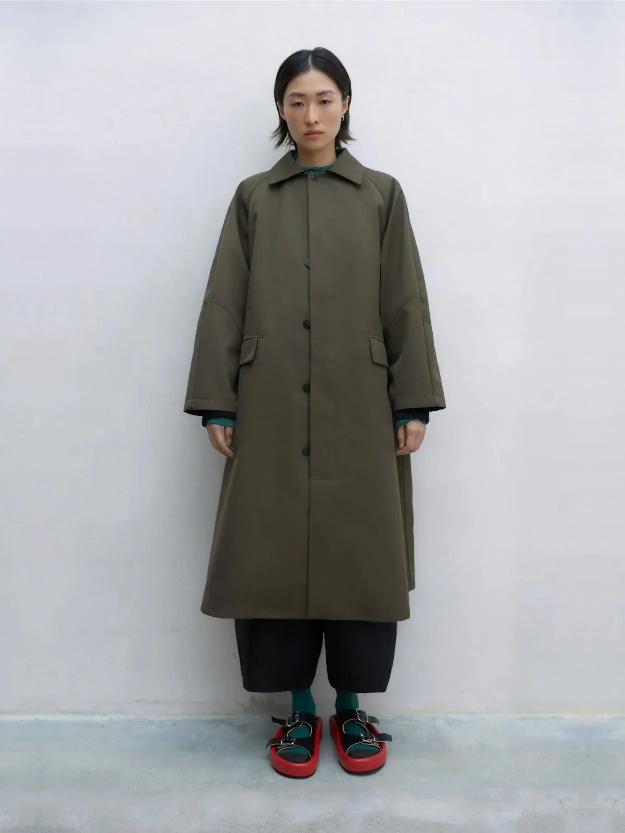 A knee-length, dark green trench coat featuring large front pockets, long sleeves, and a pointed collar. The minimalist design includes a concealed button closure and a plain back, creating a sleek and timeless look. Available at the Barcelona-based store "Bassalstore," the label "Cordera" is visible inside the collar.